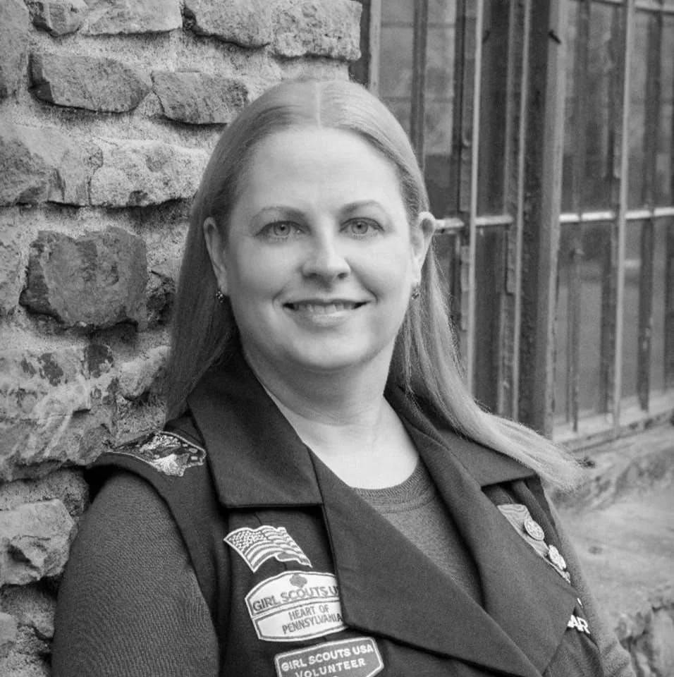 A woman in Girl Scouts uniform smiling in front of a stone wall and metal fence.