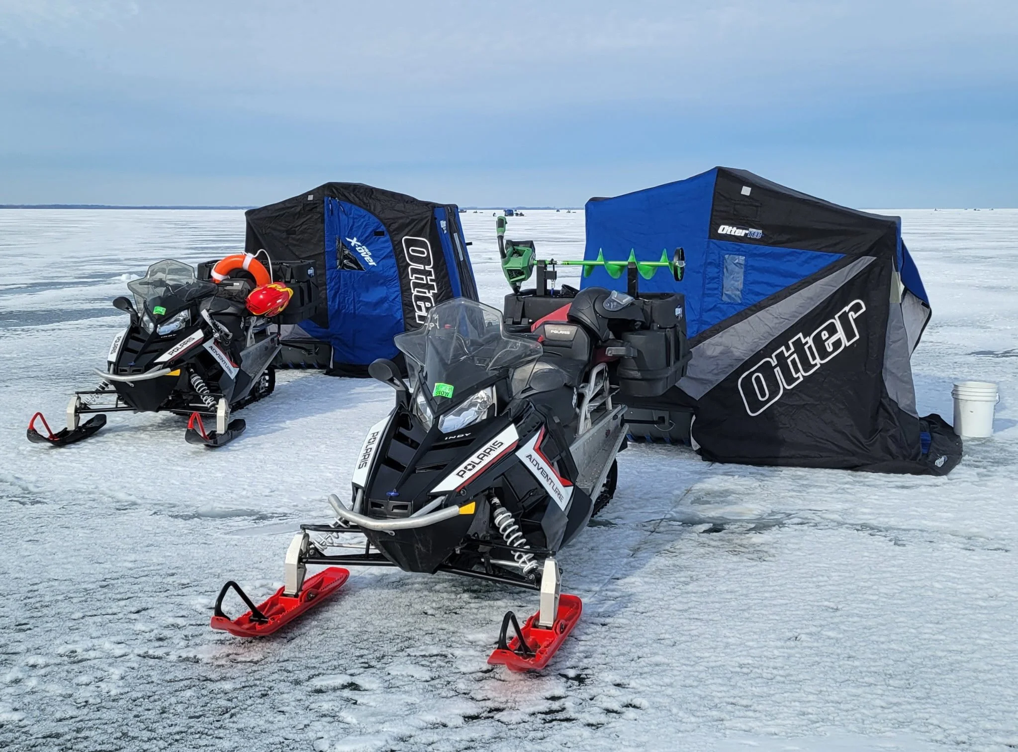Two snowmobiles and two blue-and-black ice fishing shelters sit on a frozen lake. One snowmobile carries an ice auger with green blades, along with a helmet and life preserver. The scene shows a full ice fishing setup in winter conditions.