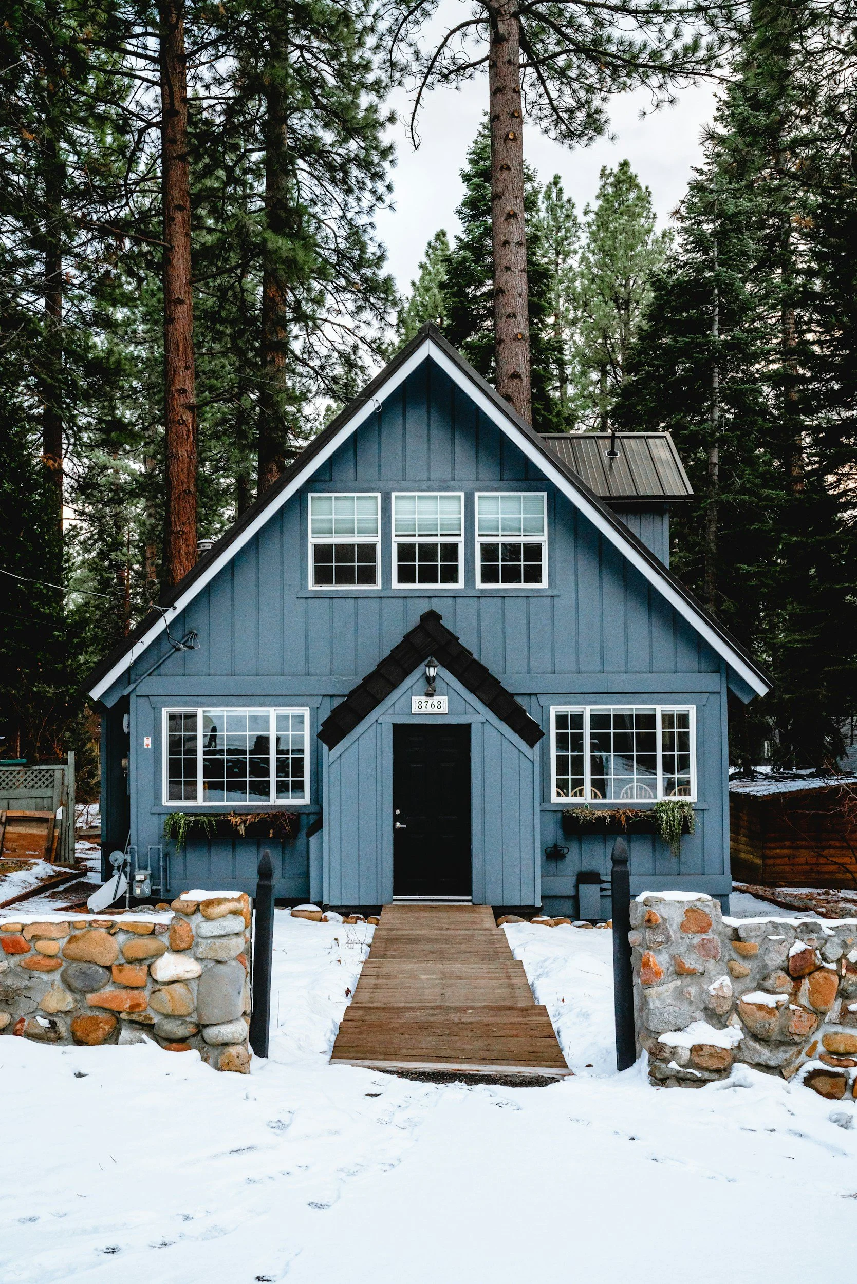 A blue A-frame cabin with large windows and a wooden path leading to the door, surrounded by snow and tall pine trees.