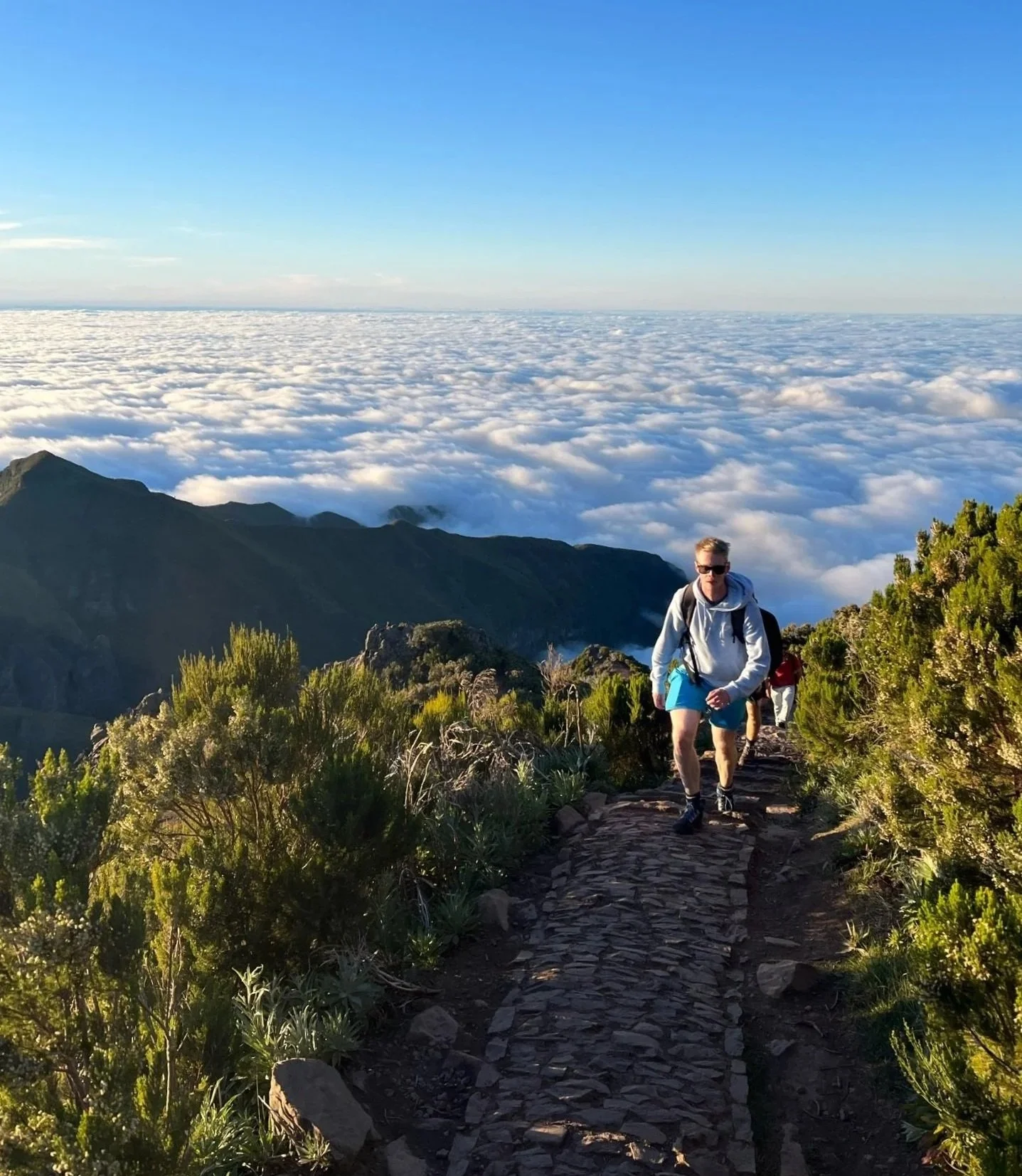 Hiker on a mountain trail with clouds and clear blue sky.