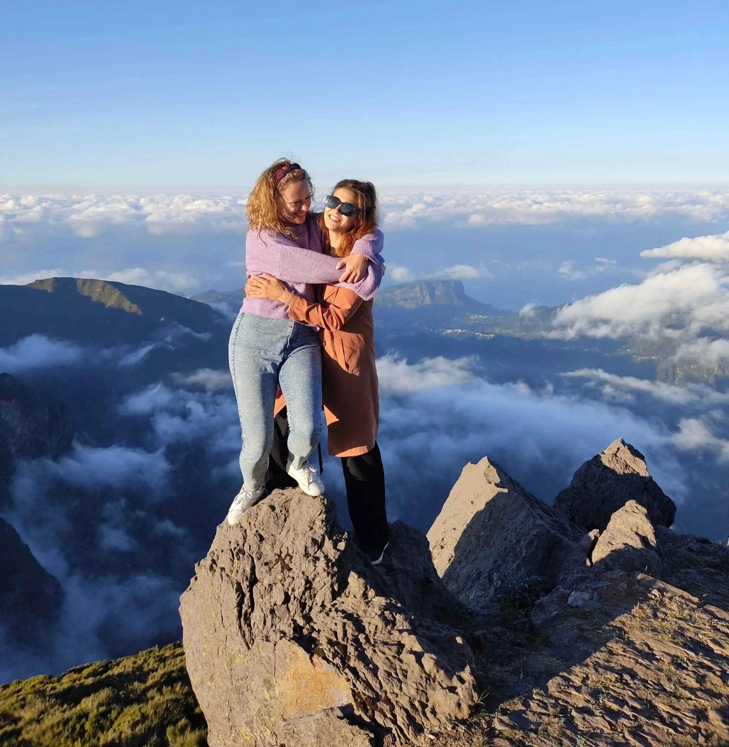 Two people standing on a mountain peak surrounded by clouds, embracing each other with a clear blue sky in the background.
