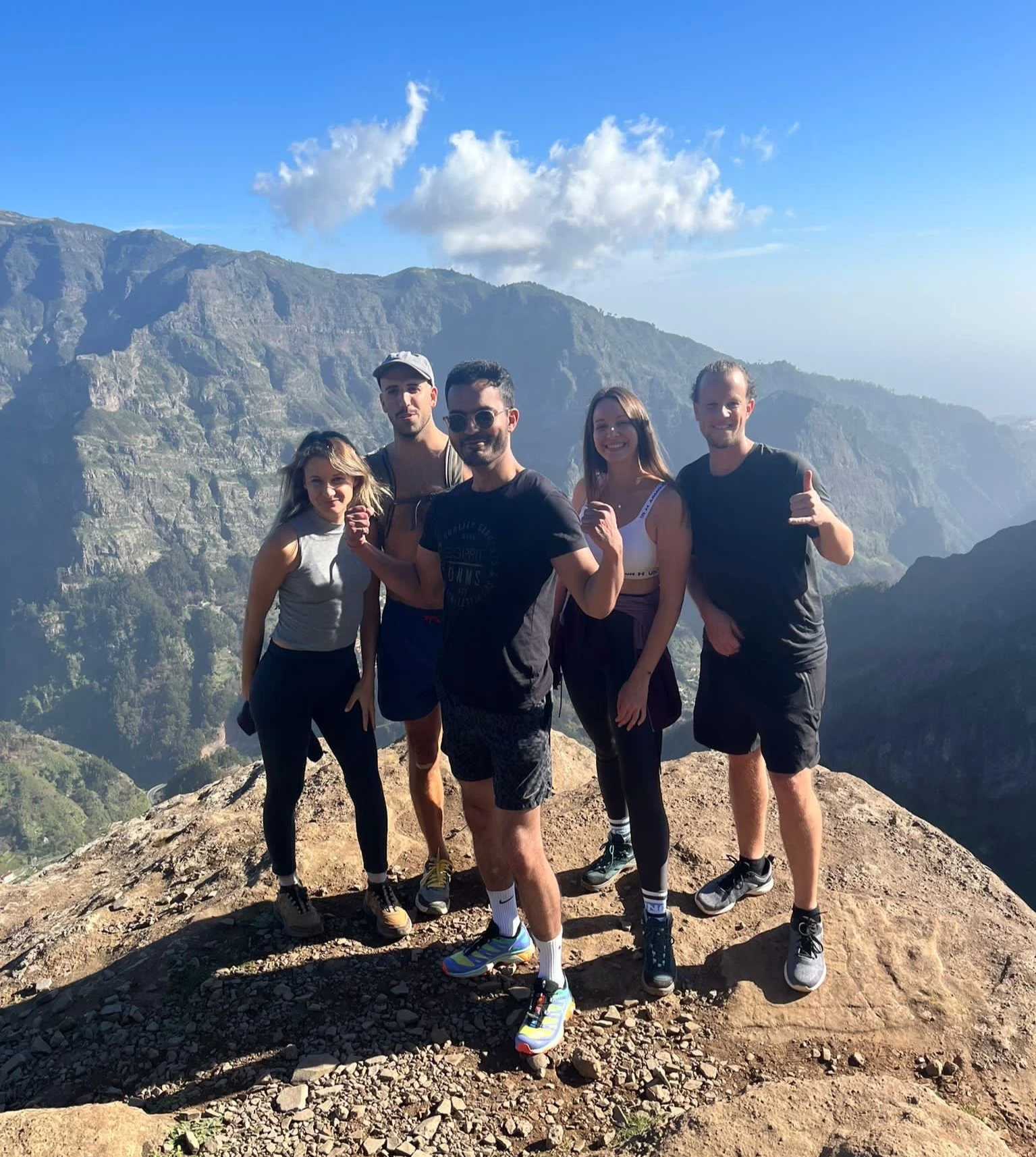 Group of five people standing on a rocky mountain peak with a scenic view of mountains and blue sky in the background