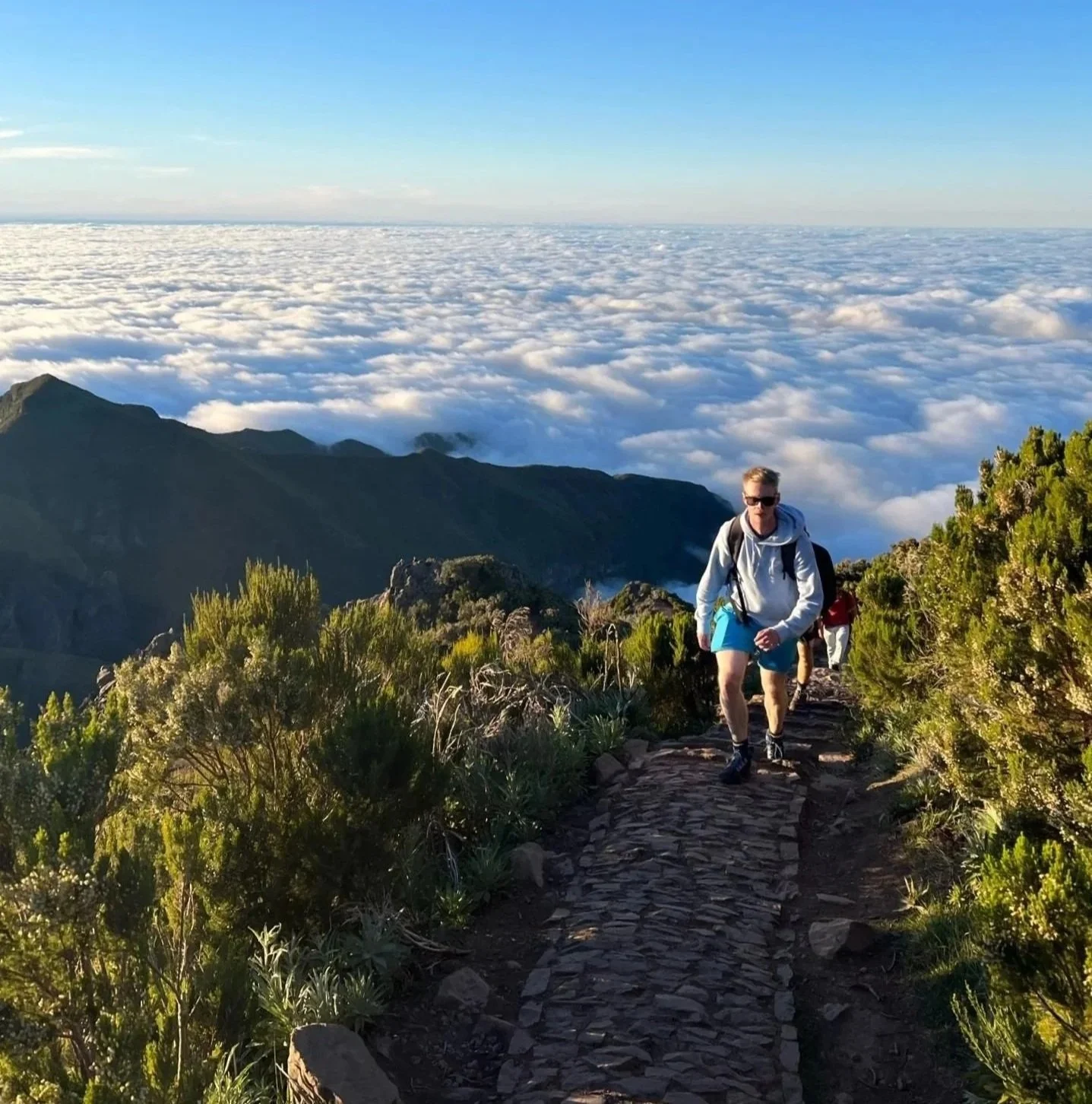 Hiker on a mountain trail with clouds and clear blue sky.