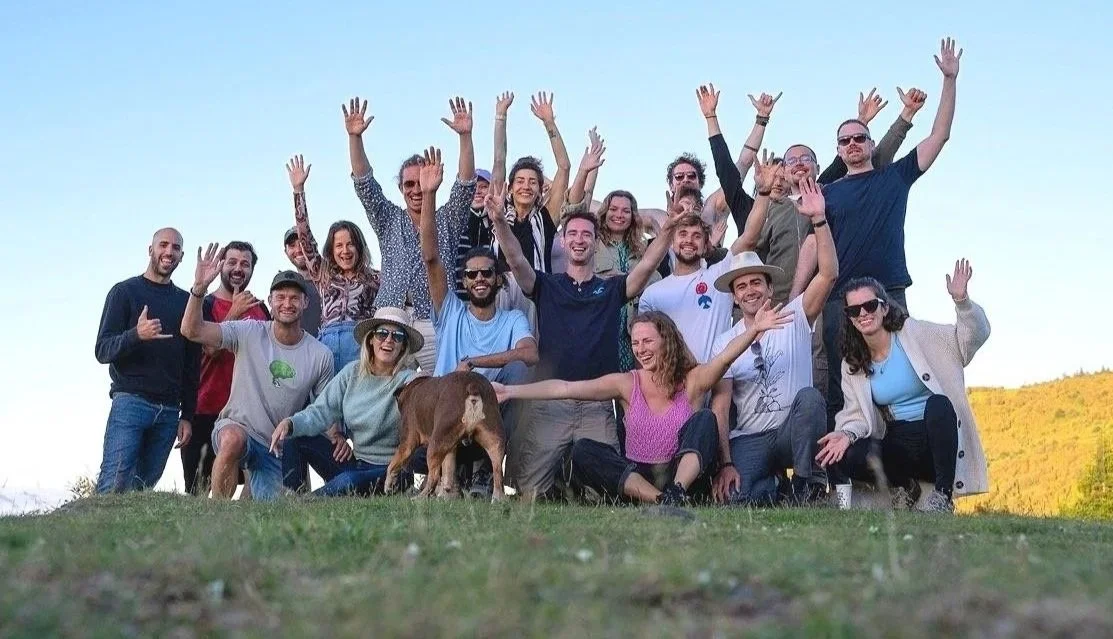 Group of 17 people outdoors smiling and raising their hands, with a dog in front, under a clear blue sky.
