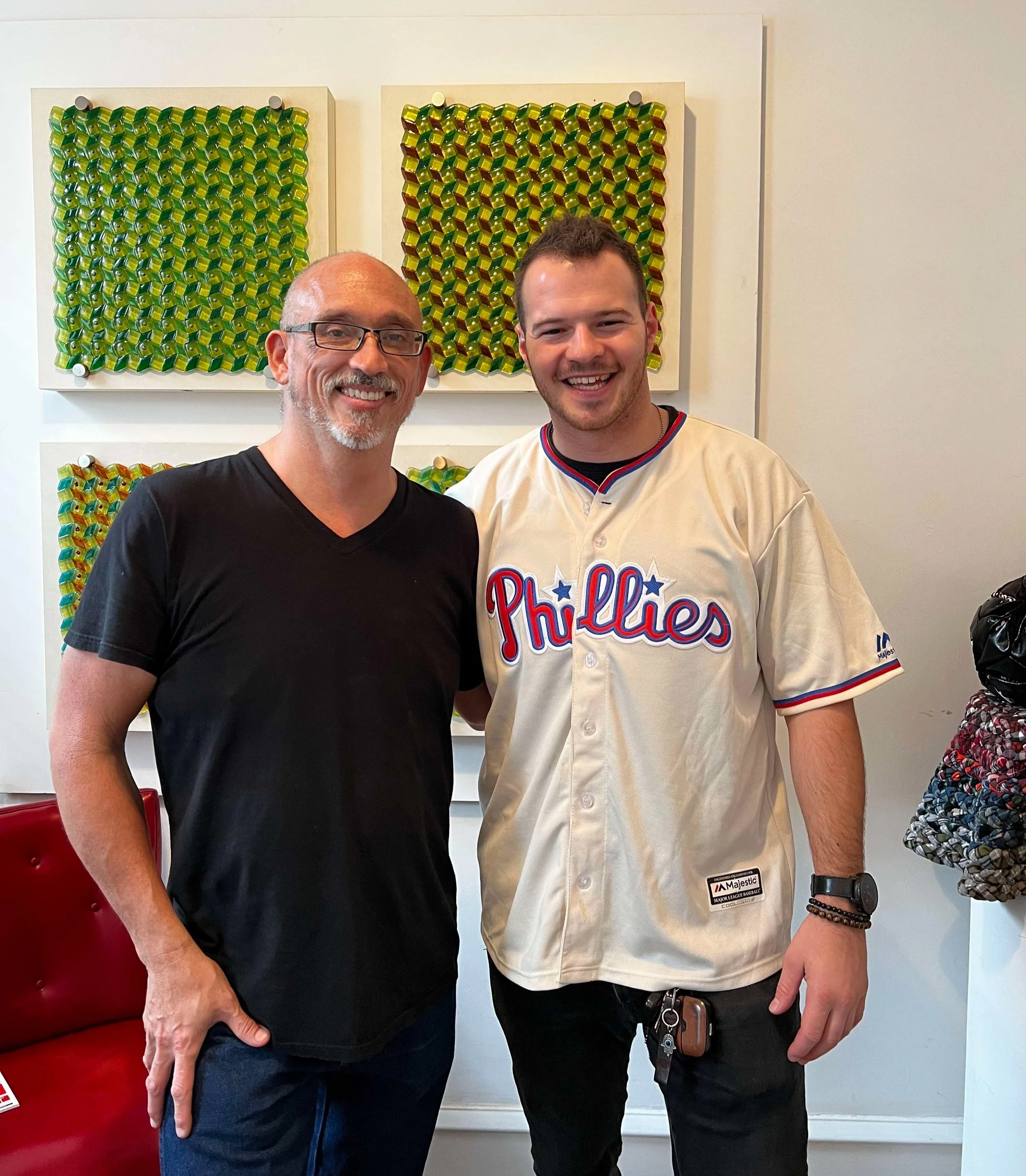 Two men smiling and standing together, one wearing a black shirt and glasses, the other wearing a Philadelphia Phillies jersey, in front of abstract artwork on the wall.