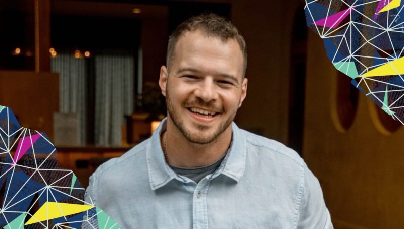 A smiling man in a light blue shirt standing indoors with wooden decor and colorful geometric patterns in the corners.