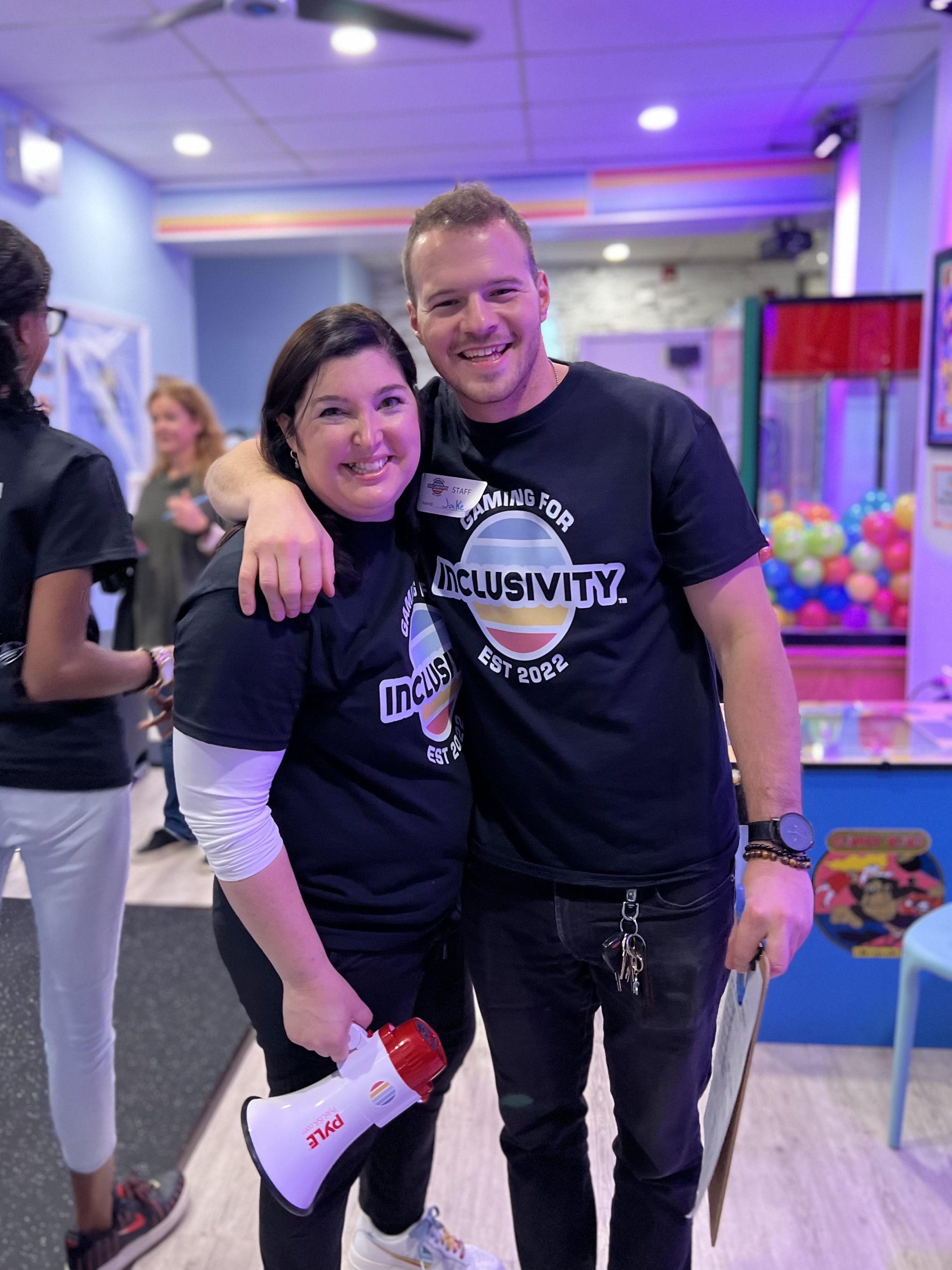 A smiling woman and man in matching black t-shirts with the logo 'Gaming for Inclusivity' pose together, with the man holding a megaphone, at what appears to be an indoor arcade or game center.
