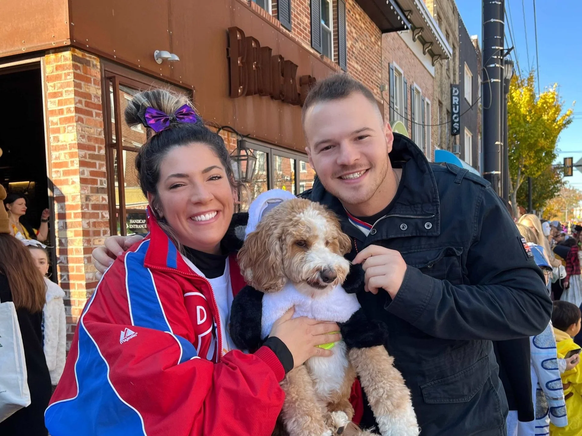 Smiling woman and man holding a puppy in front of a brick building during a street event, with a crowd in the background.