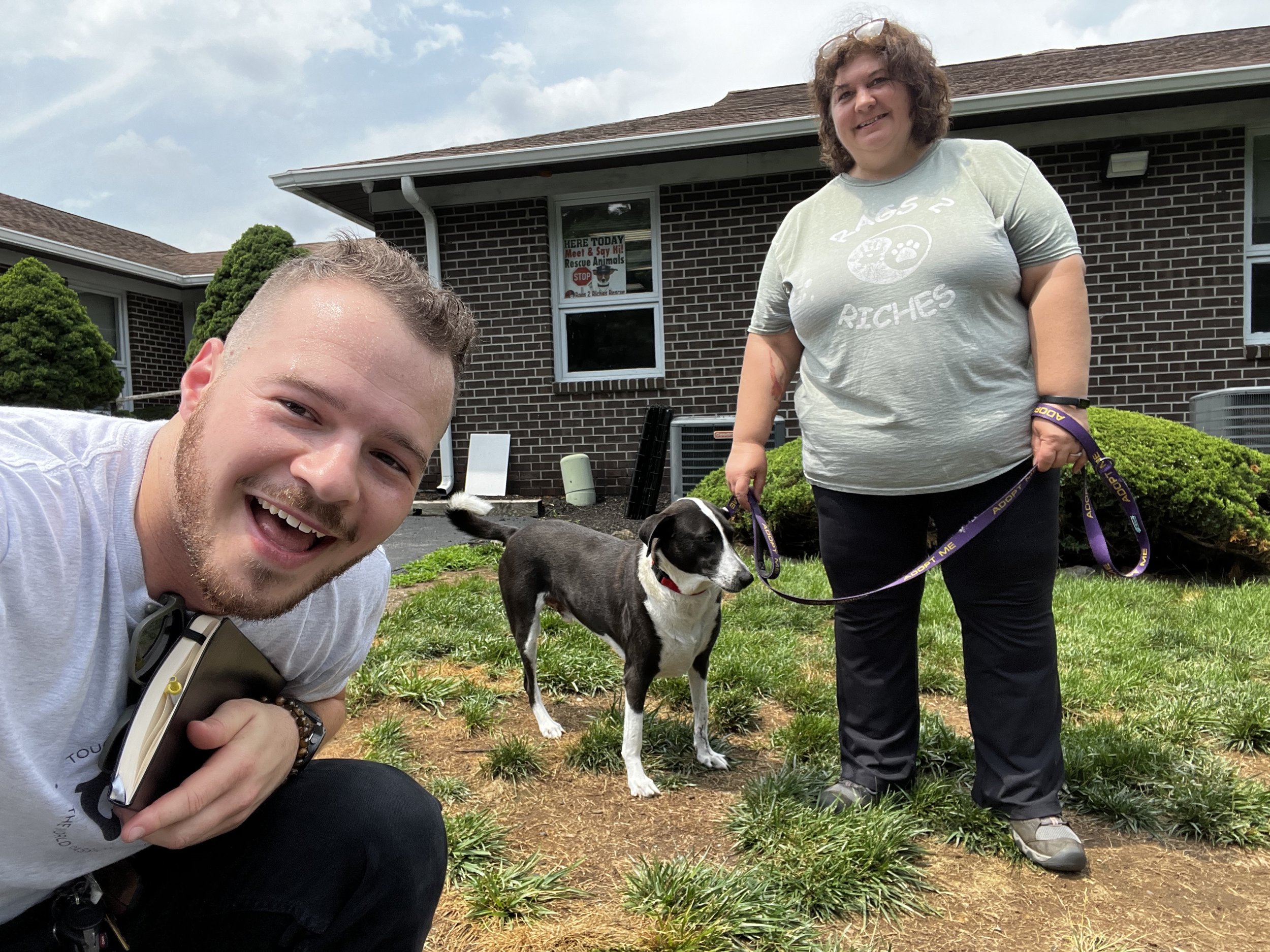 A smiling young man taking a selfie with a woman holding a leash attached to a black and white dog. They are outside in front of a brick house with bushes, on a grassy patch.