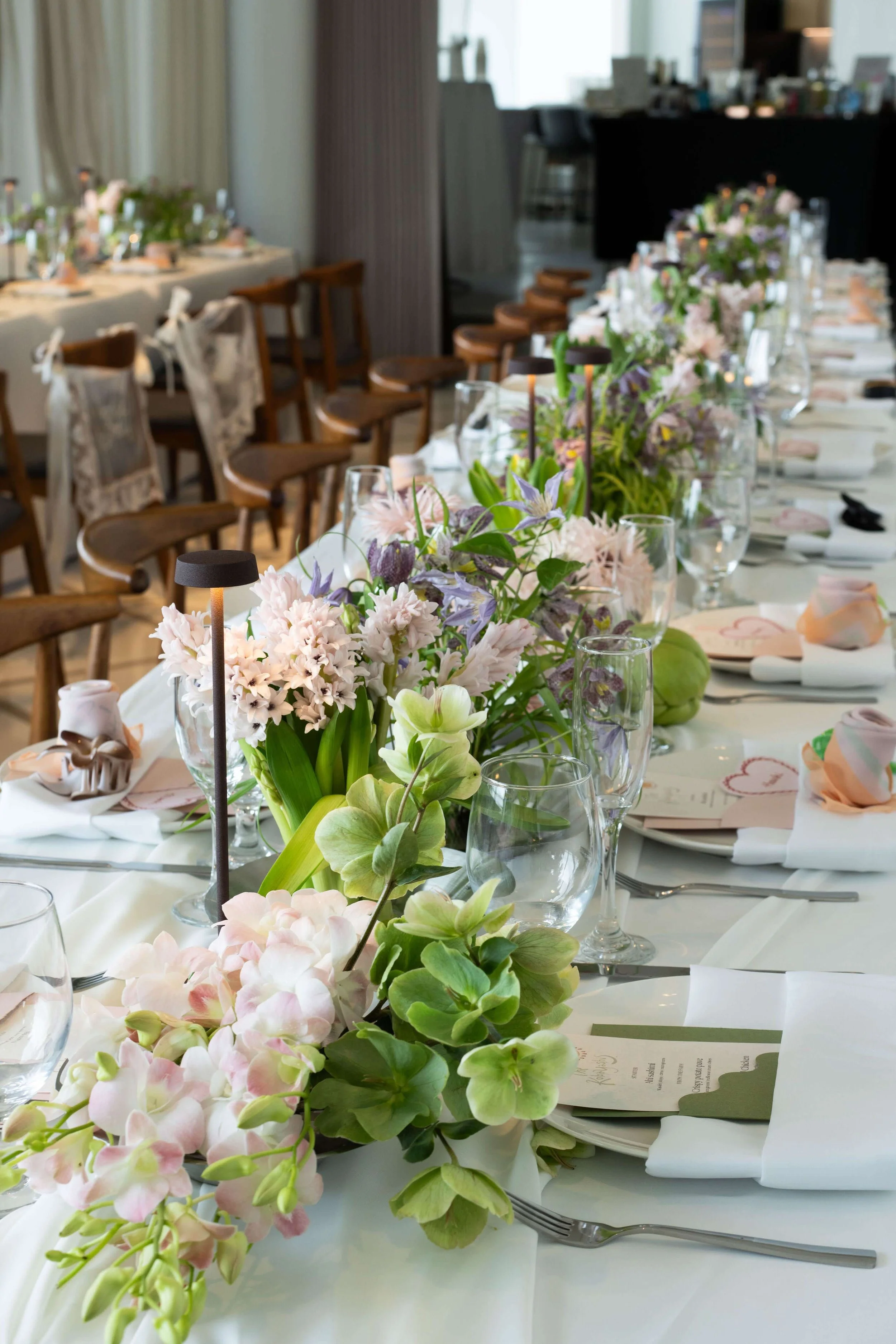 Decorated dining table with floral centerpieces, wine glasses, napkins, and place cards in a well-lit room.