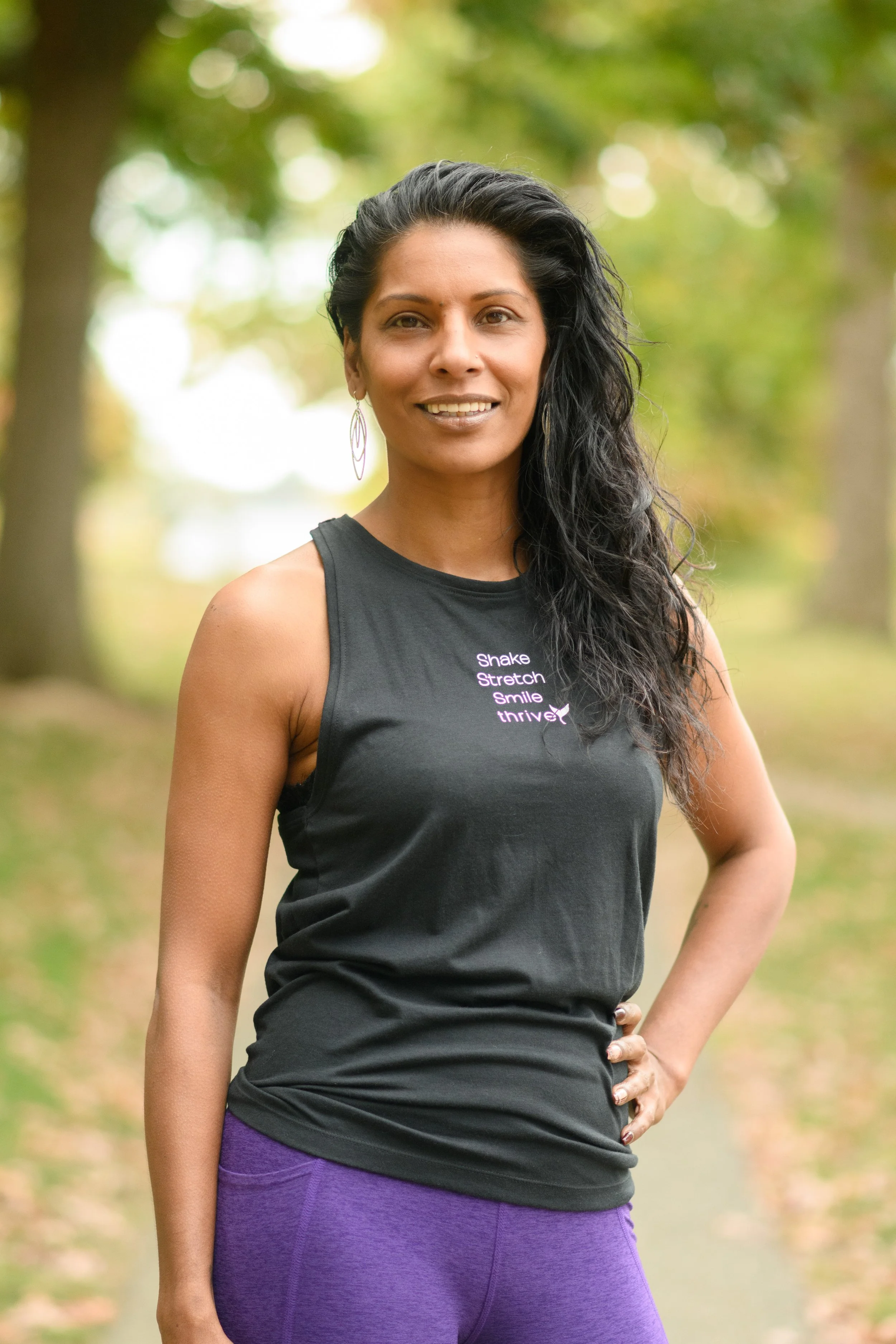 Woman smiling outdoors, wearing a black tank top with "Shake, Stretch, Smile, Thrive" text and purple leggings.