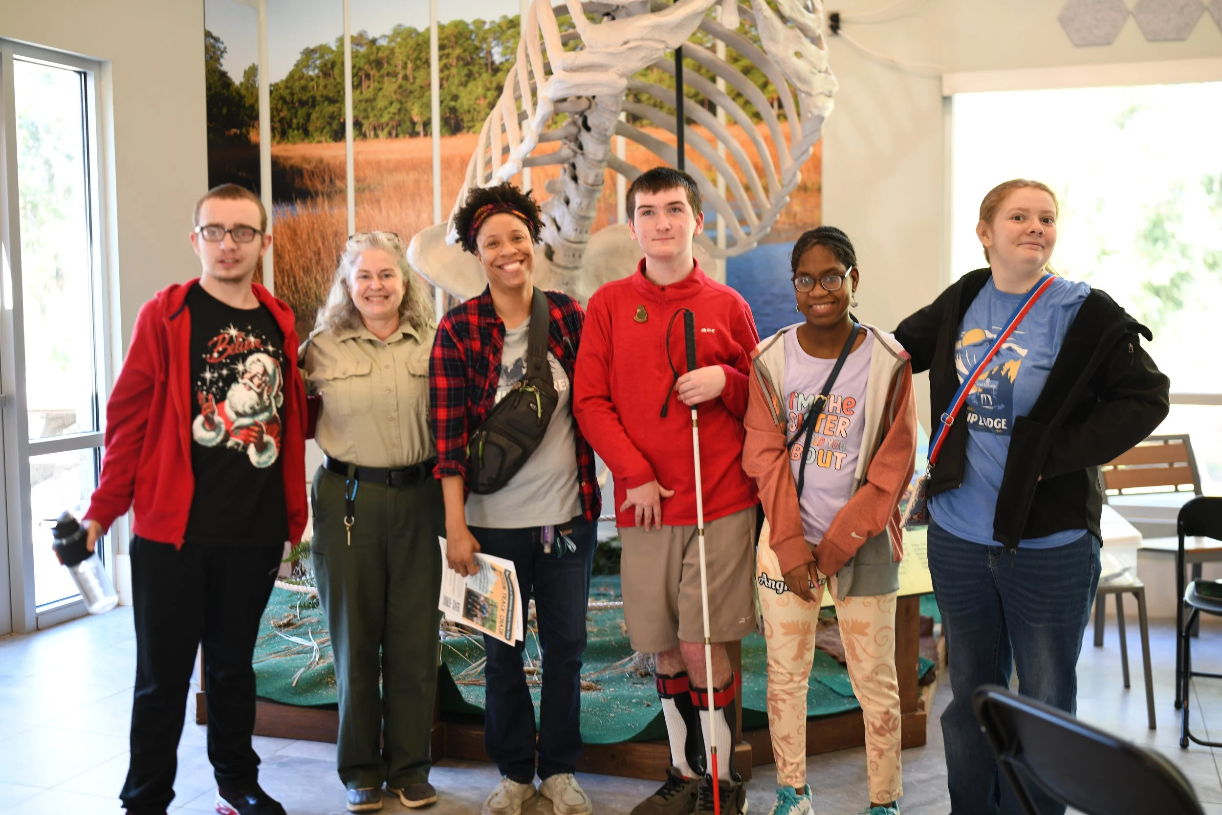 Image shows a group of students with disabilities standing with their instructor and a Park Ranger standing in front of a 20-foot tall replica skeleton of an extinct giant ground sloth