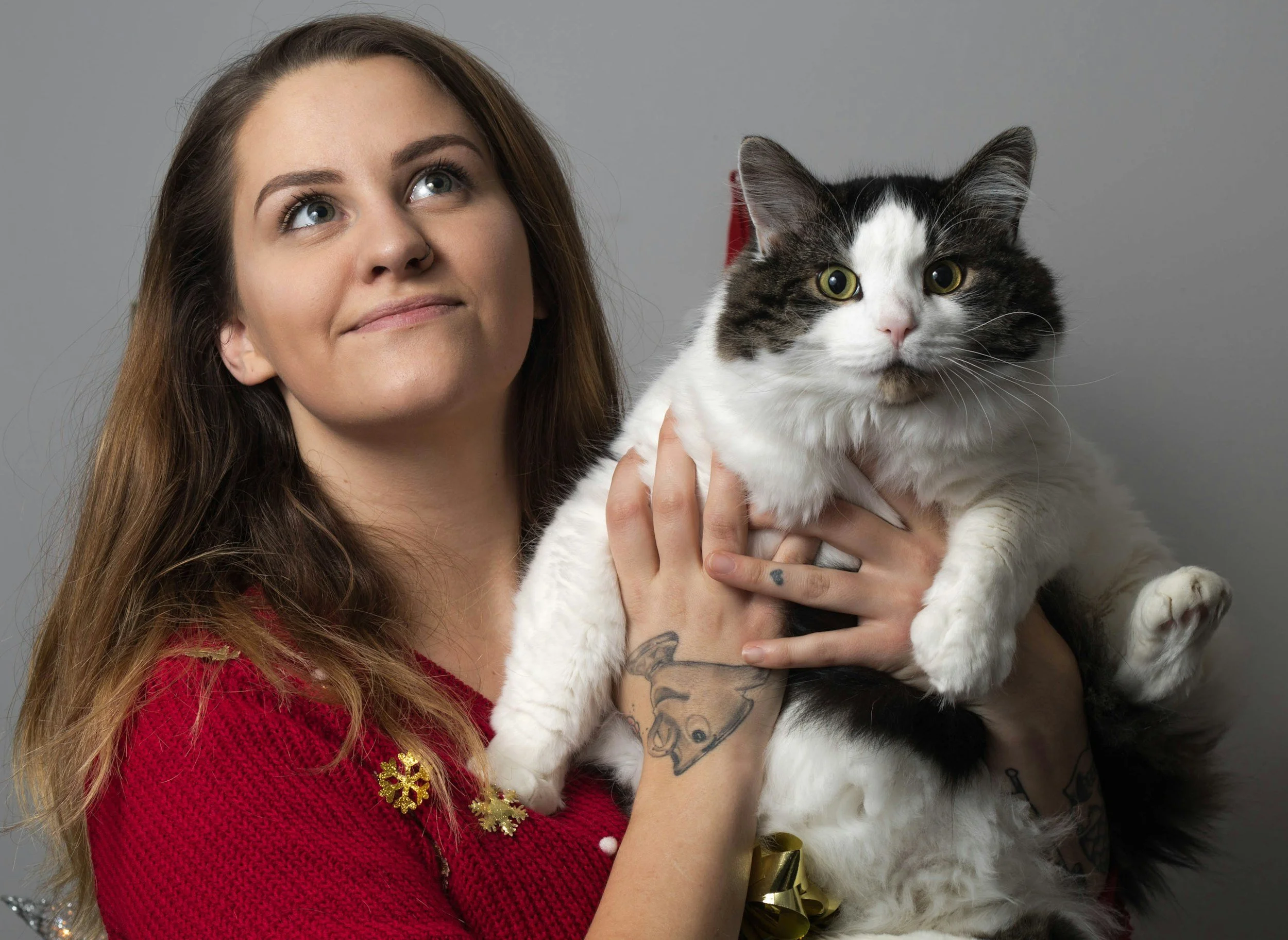 A woman with long brown hair holding a large black and white cat, both looking at the camera, against a plain gray background.