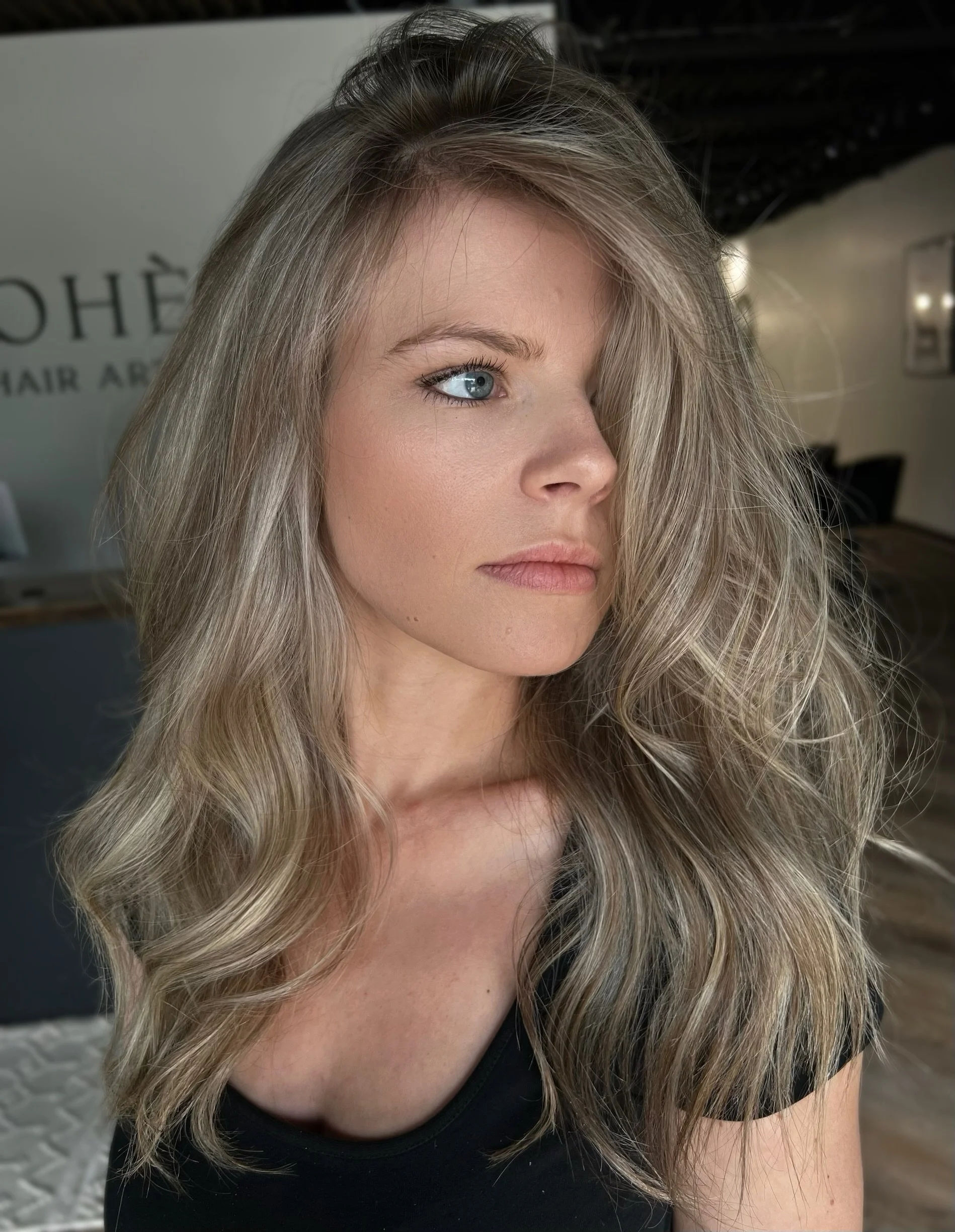 Close-up of a woman with long, wavy blonde hair, wearing a black top, looking to the side, with a blurred background that includes a wall with some text and part of a salon or studio interior.