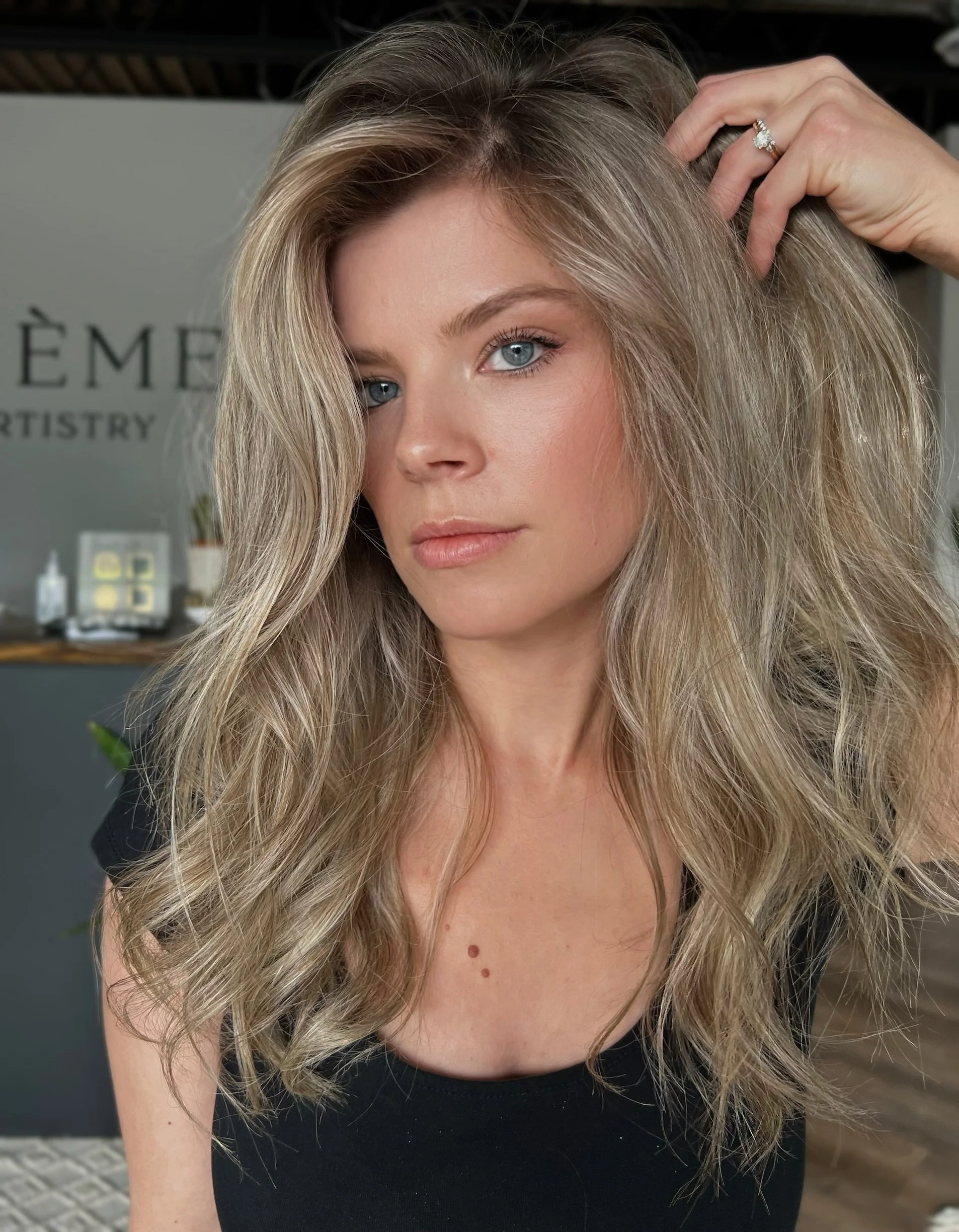 A woman with long, wavy blonde hair, blue eyes, and fair skin, smiling slightly while touching her hair, wearing a silver ring.