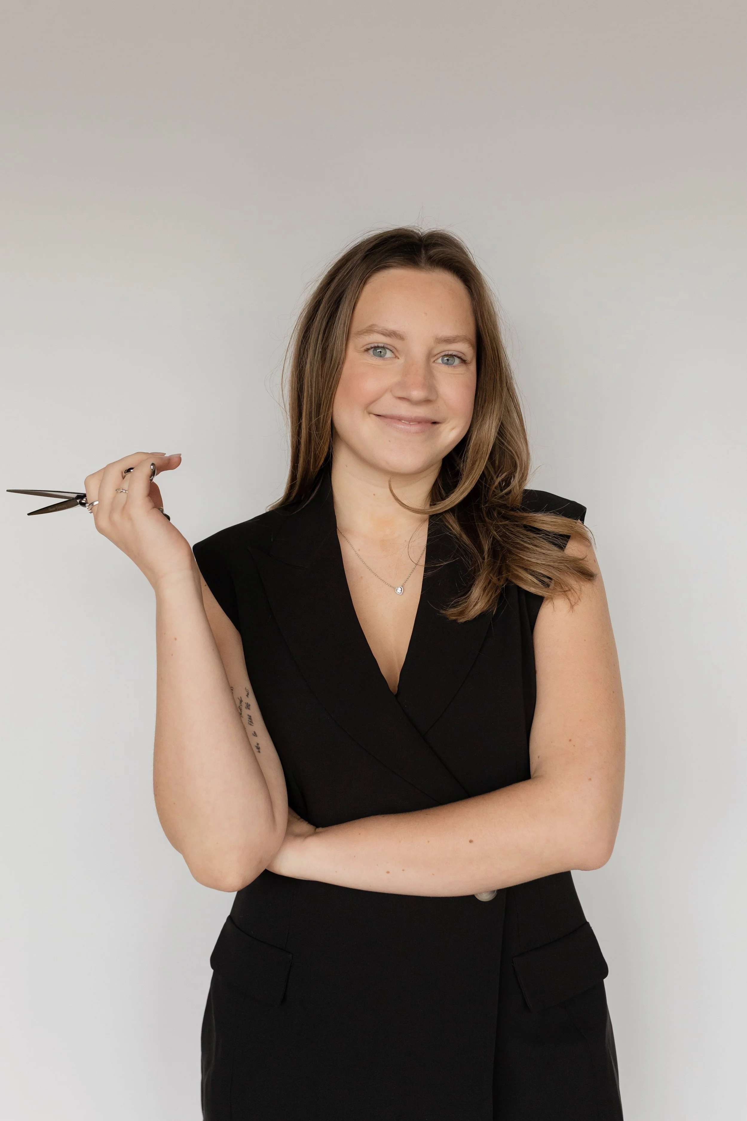 A young woman with long brown hair and blue eyes wearing a sleeveless black dress, holding scissors in her right hand, smiling, standing against a plain light gray background.