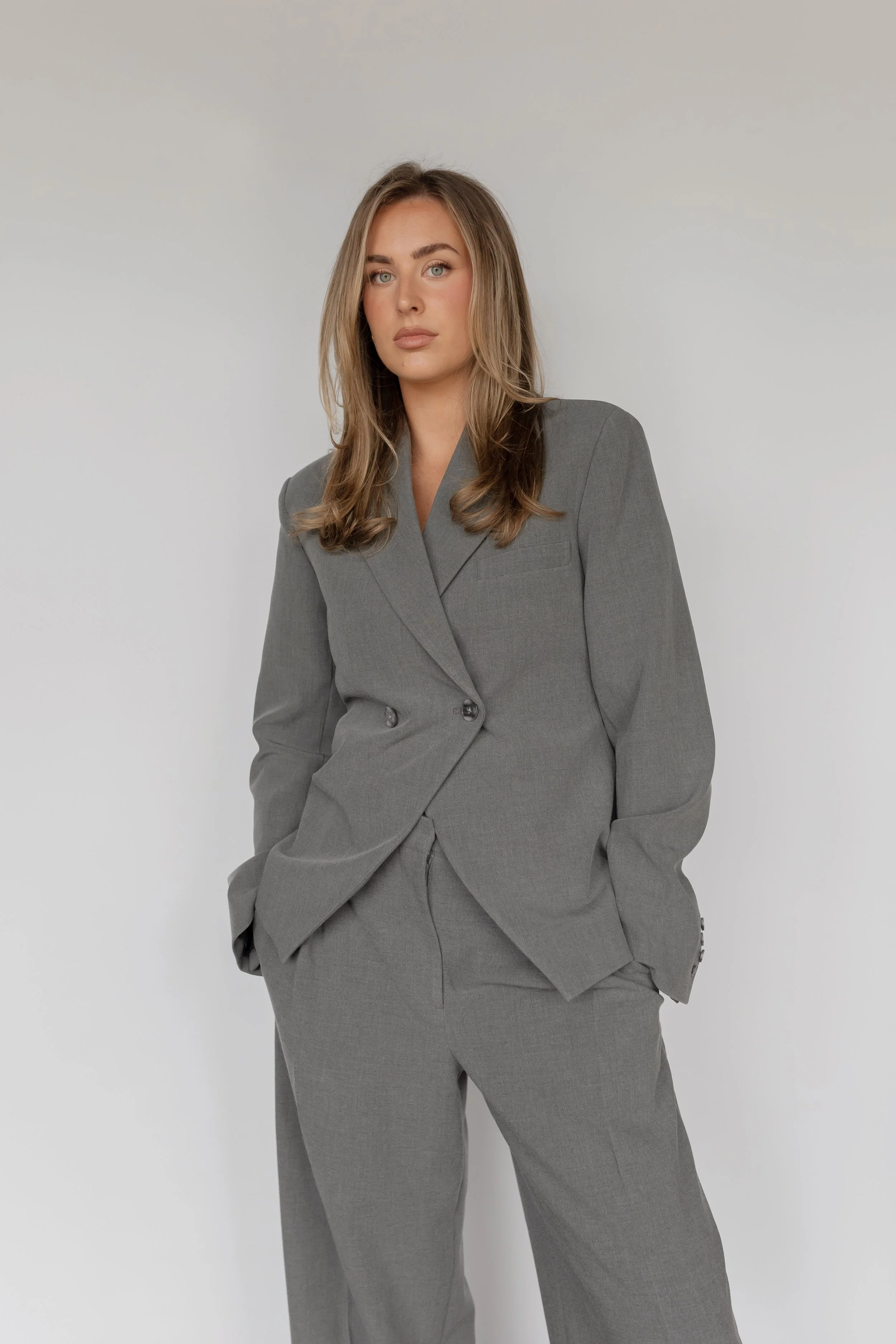 A young woman with long, wavy blonde hair and blue eyes wearing a gray business suit with her left hand in her pocket, standing against a plain white background.