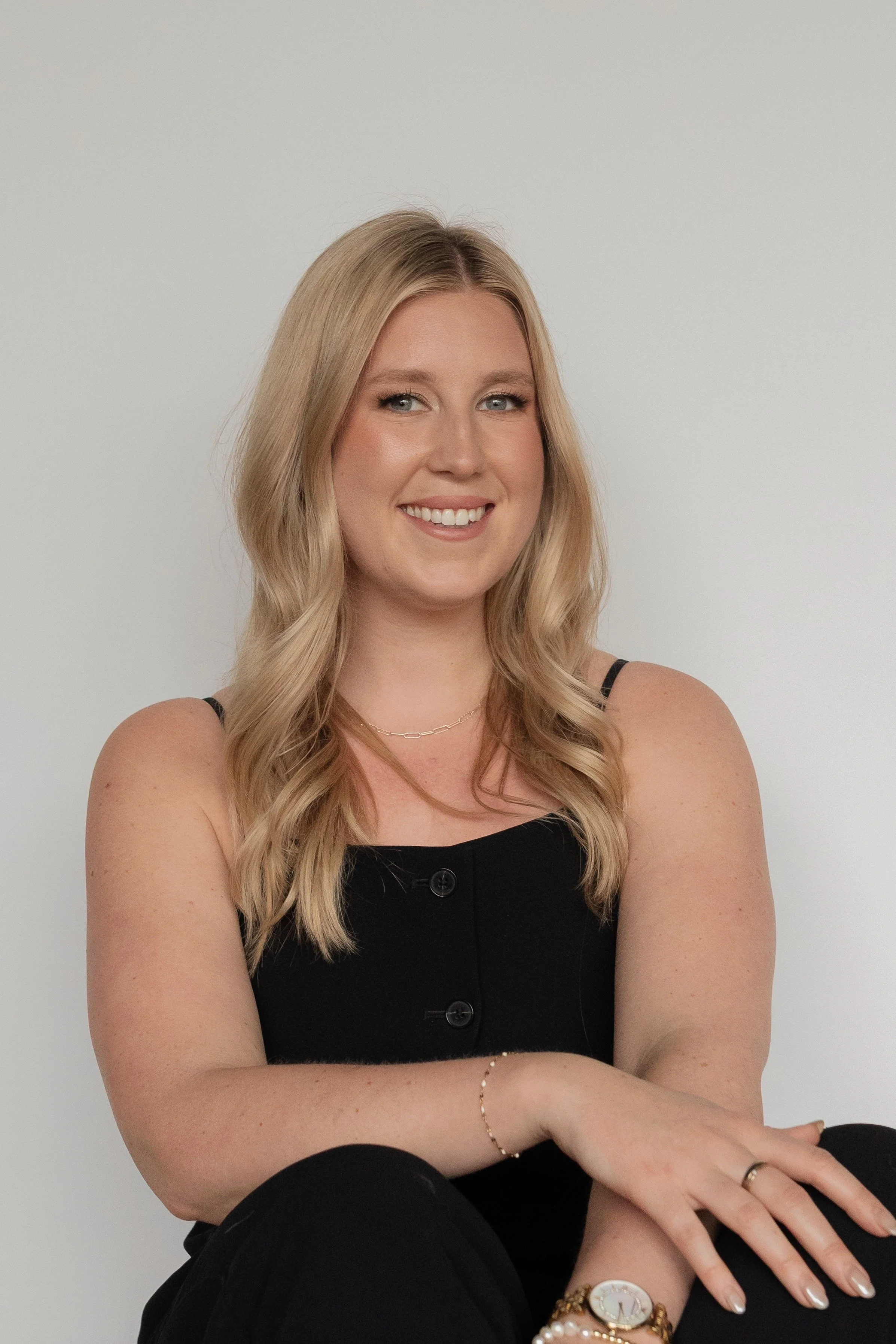 Portrait of a smiling young woman with long blonde hair, wearing a black sleeveless top, jewelry including a watch, bracelets, a necklace, and rings, sitting with crossed arms against a plain light gray background.