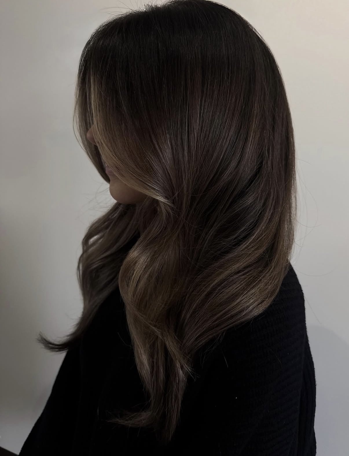 A woman with long, wavy hair with dark brown roots and lighter brown ends, wearing a black top, standing against a plain light-colored background.