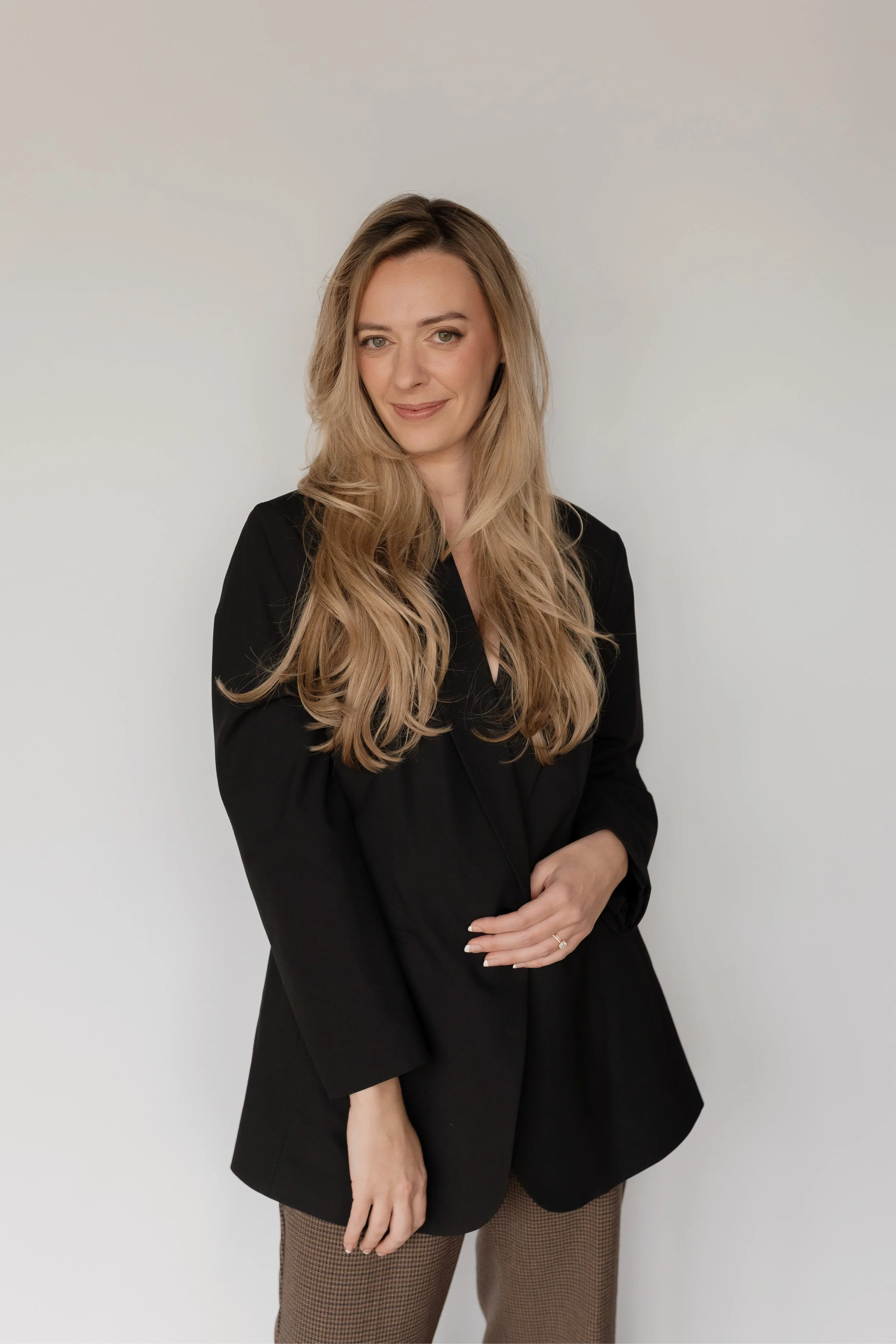 A woman with long wavy blonde hair wearing a black blazer, black dress, and checkered pants standing against a plain white background.
