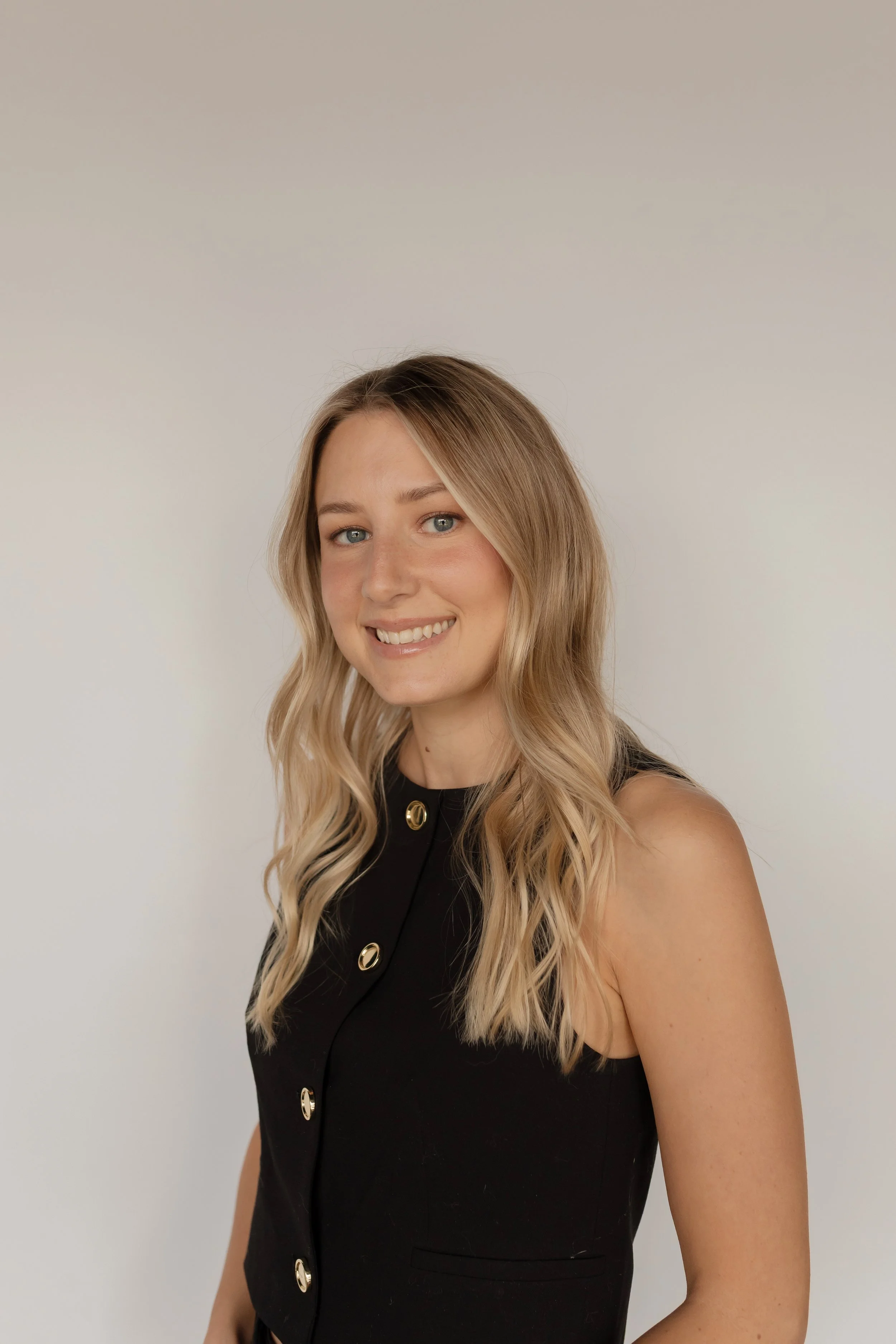 A young woman with long blonde wavy hair taking a selfie in front of a plain off-white wall. She is smiling and wearing a black sleeveless dress with gold buttons.