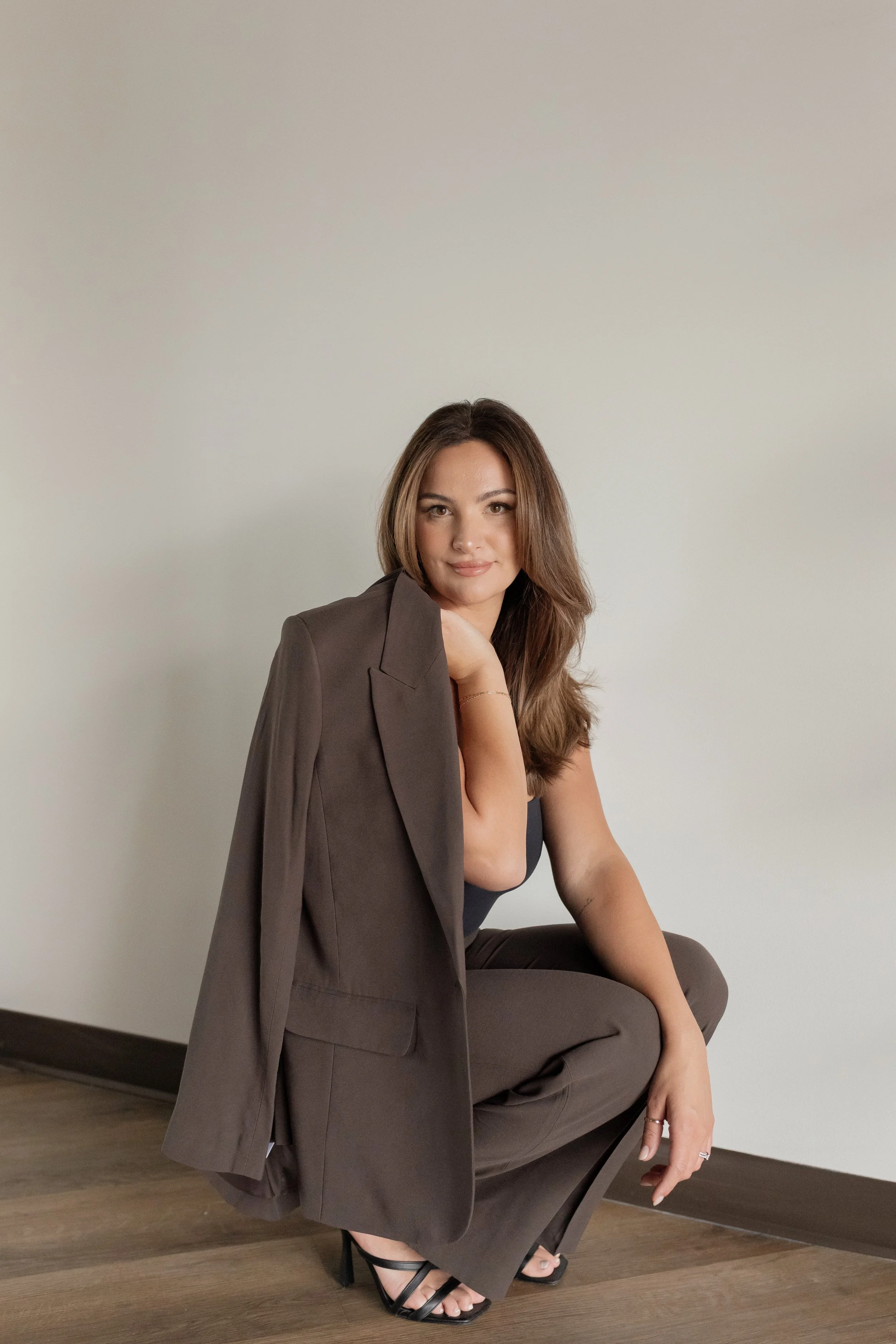 A woman with long brown hair sitting on a chair with a beige wall background, wearing a dark-colored outfit and black high-heeled sandals, with a blazer draped over her shoulder.