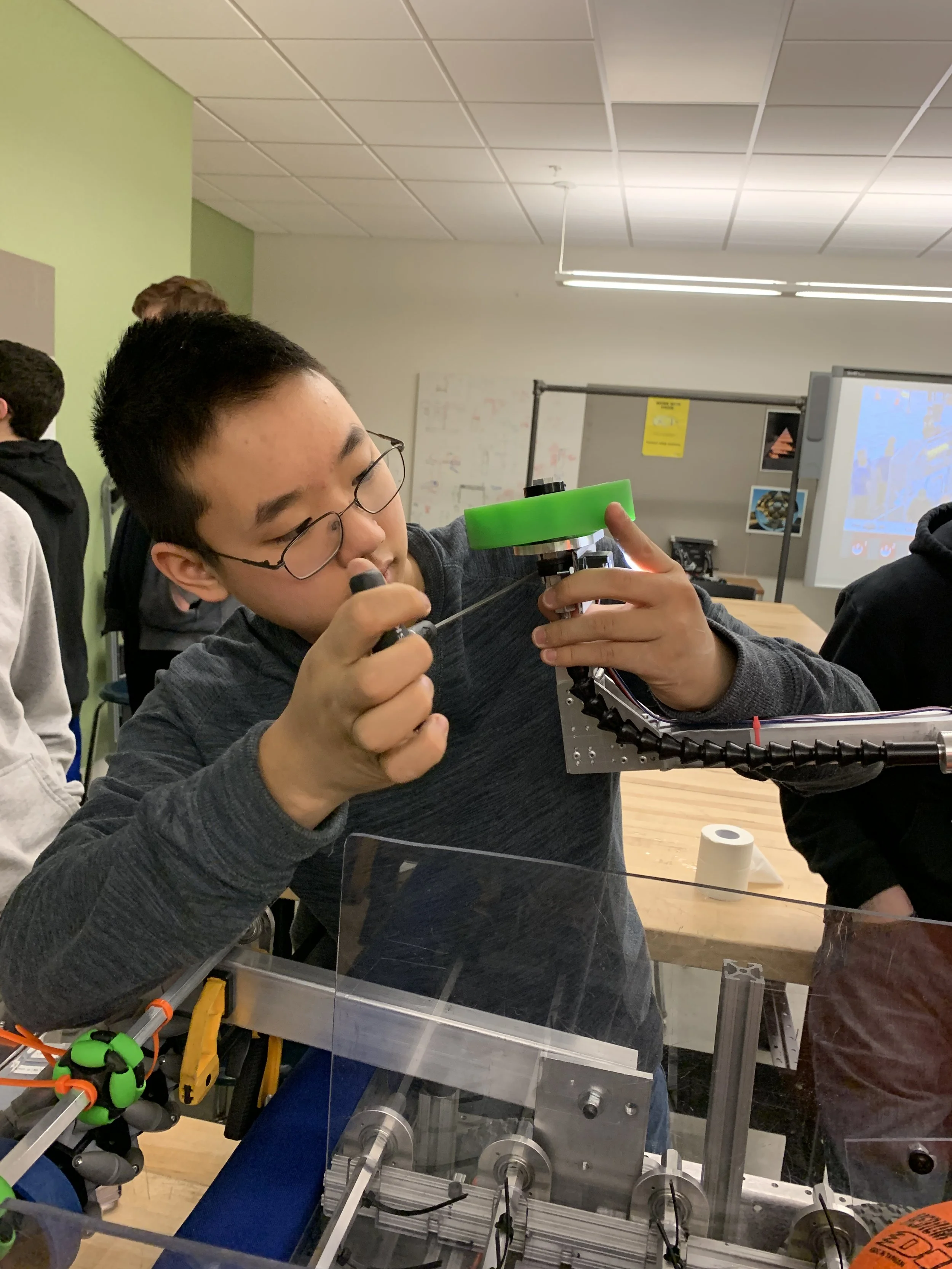 A young person with glasses working on a mechanical device or robot, adjusting components with a screwdriver in a classroom or lab setting.