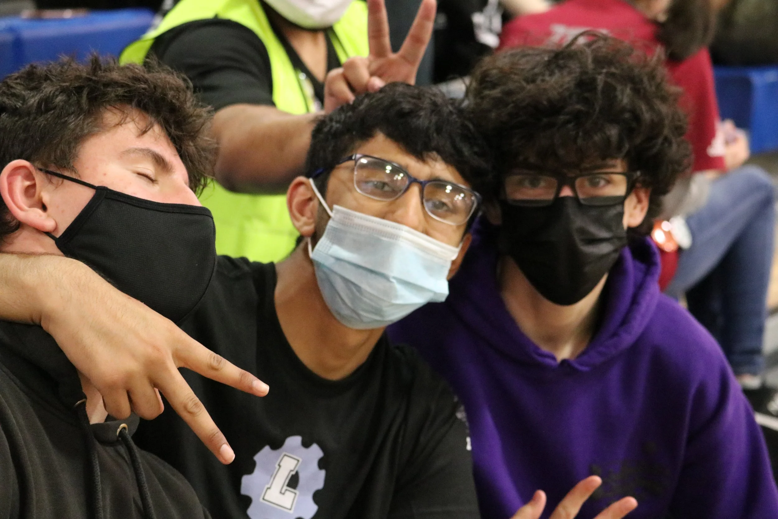Three young males wearing face masks, posing for a photo, with a woman in the background making a peace sign.