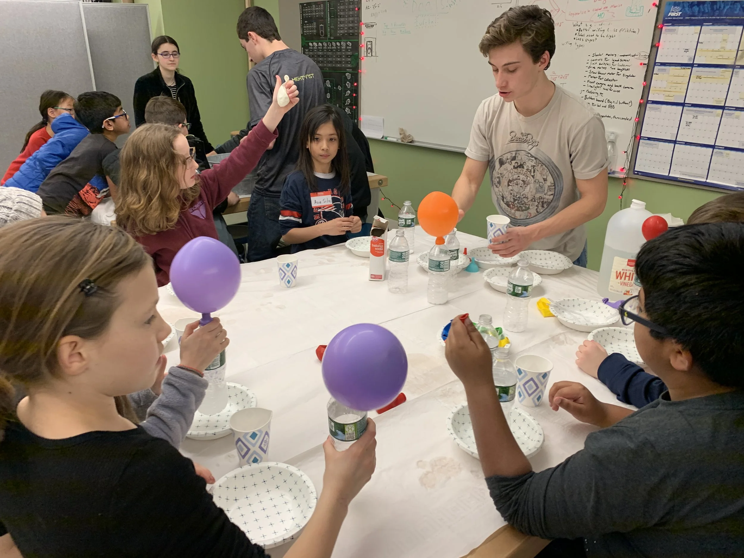 Children gathered around a table with balloons, paper plates, and water bottles, with a young man showing a science demonstration involving a balloon on top of a plastic bottle.