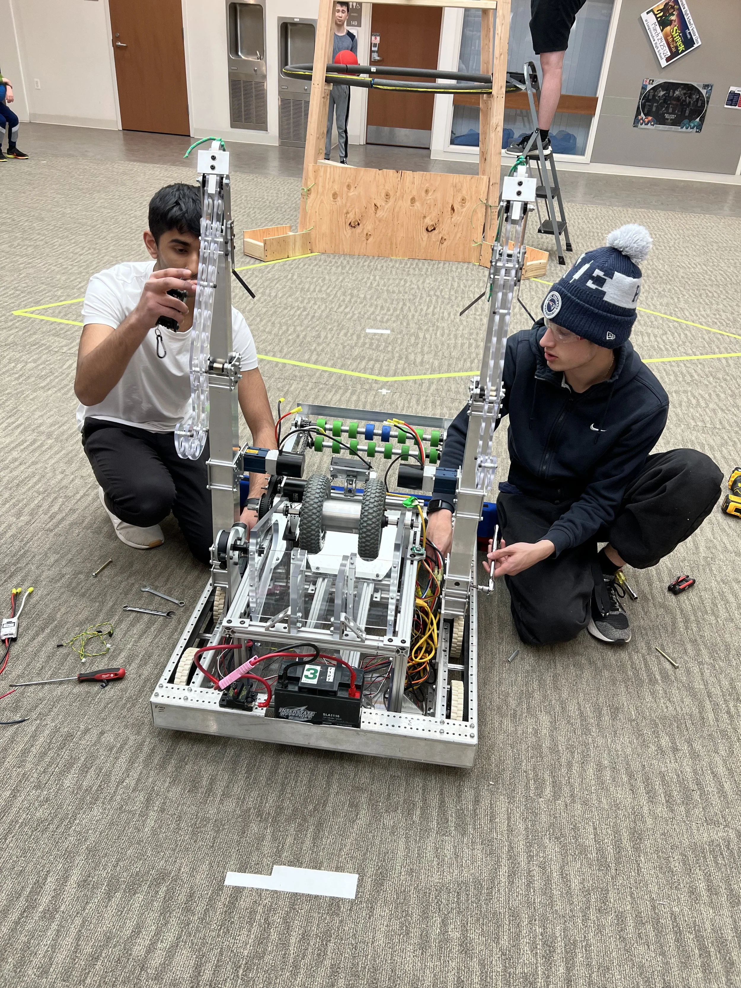 Two young men working on a robot in a room, surrounded by tools and wires, with other children and objects in the background.