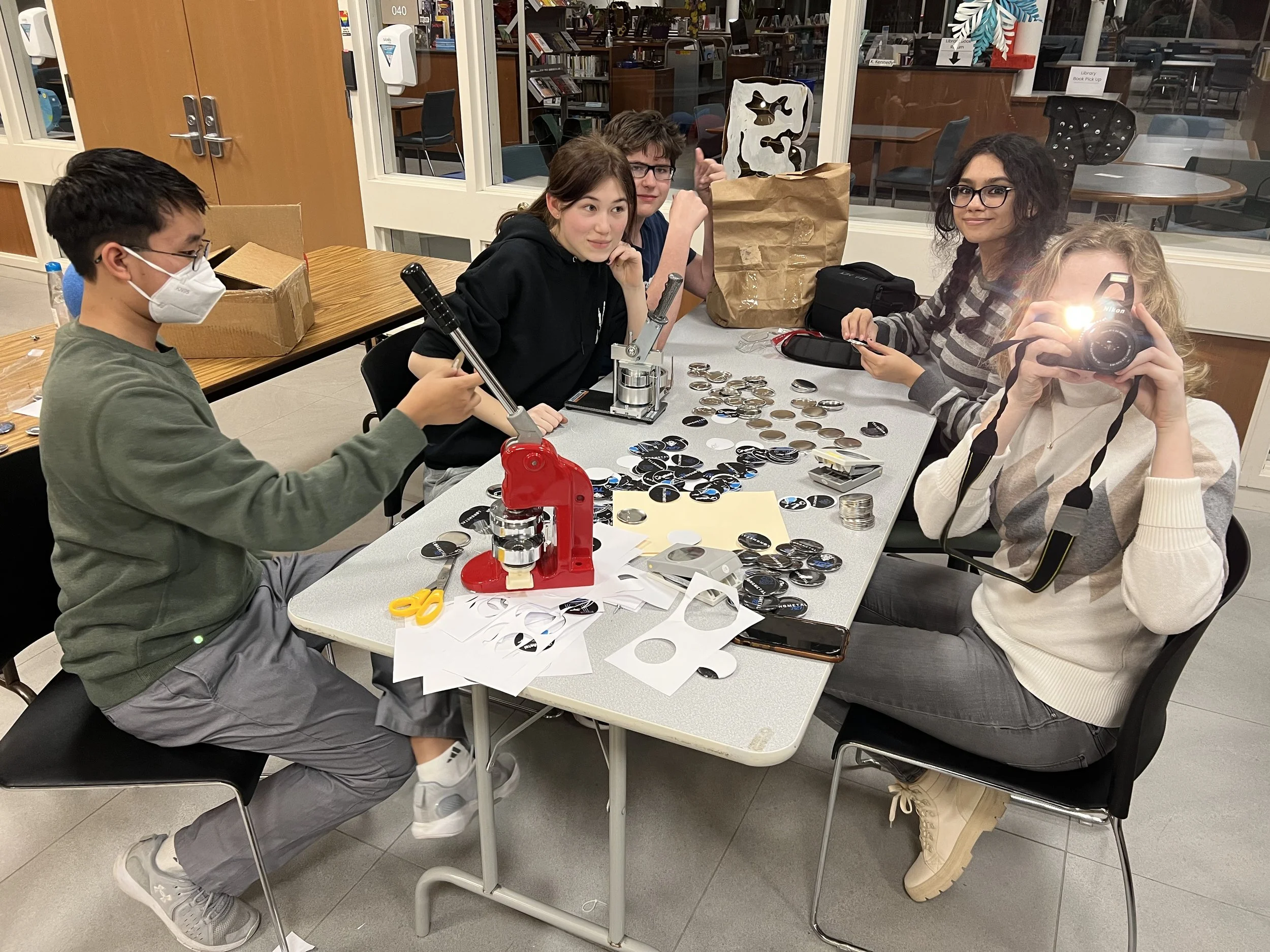 A group of five young people working on sorting and possibly assembling buttons or badges at a table. They are in a room with glass windows, shelves, and tables in the background. One person is wearing a face mask and using a tool, another is taking 