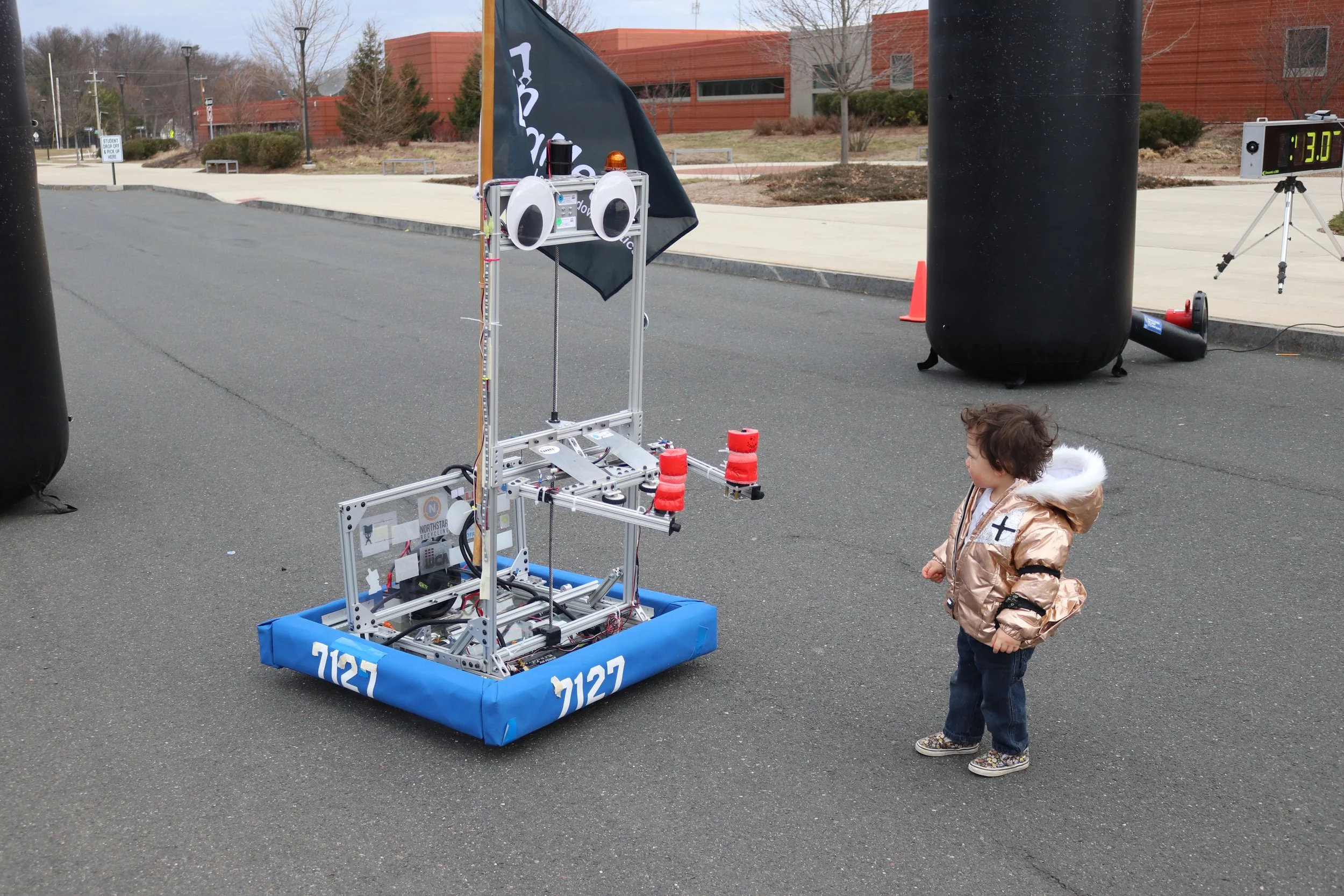 A young child in a gold jacket watching a robot competition on an outdoor street, with a robot and a digital timer visible in the background.