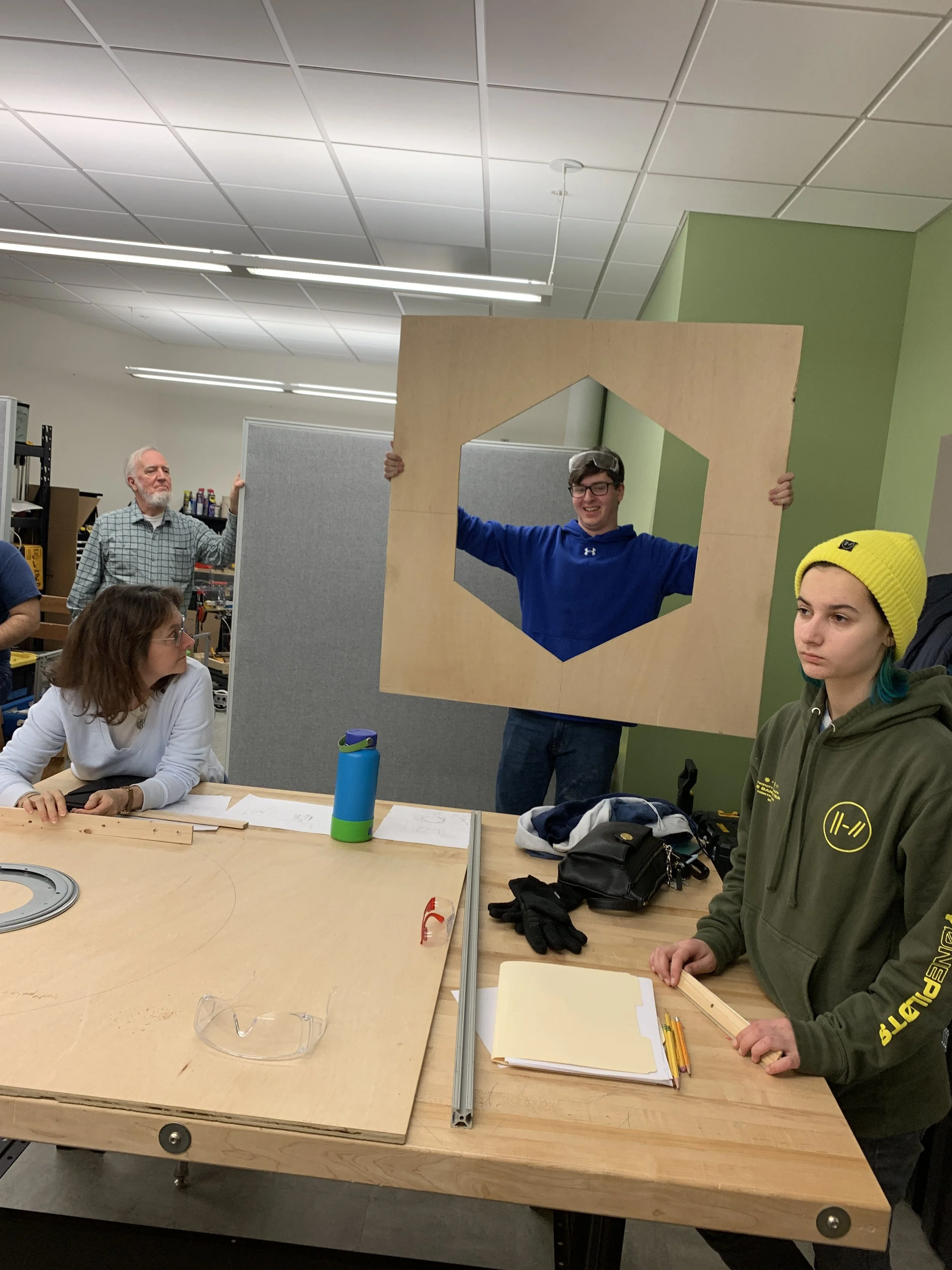 A group of young and older people in a workshop or classroom setting, with some holding a large wooden frame with a hexagon-shaped cutout, while others sit at a table with tools, papers, and a water bottle.