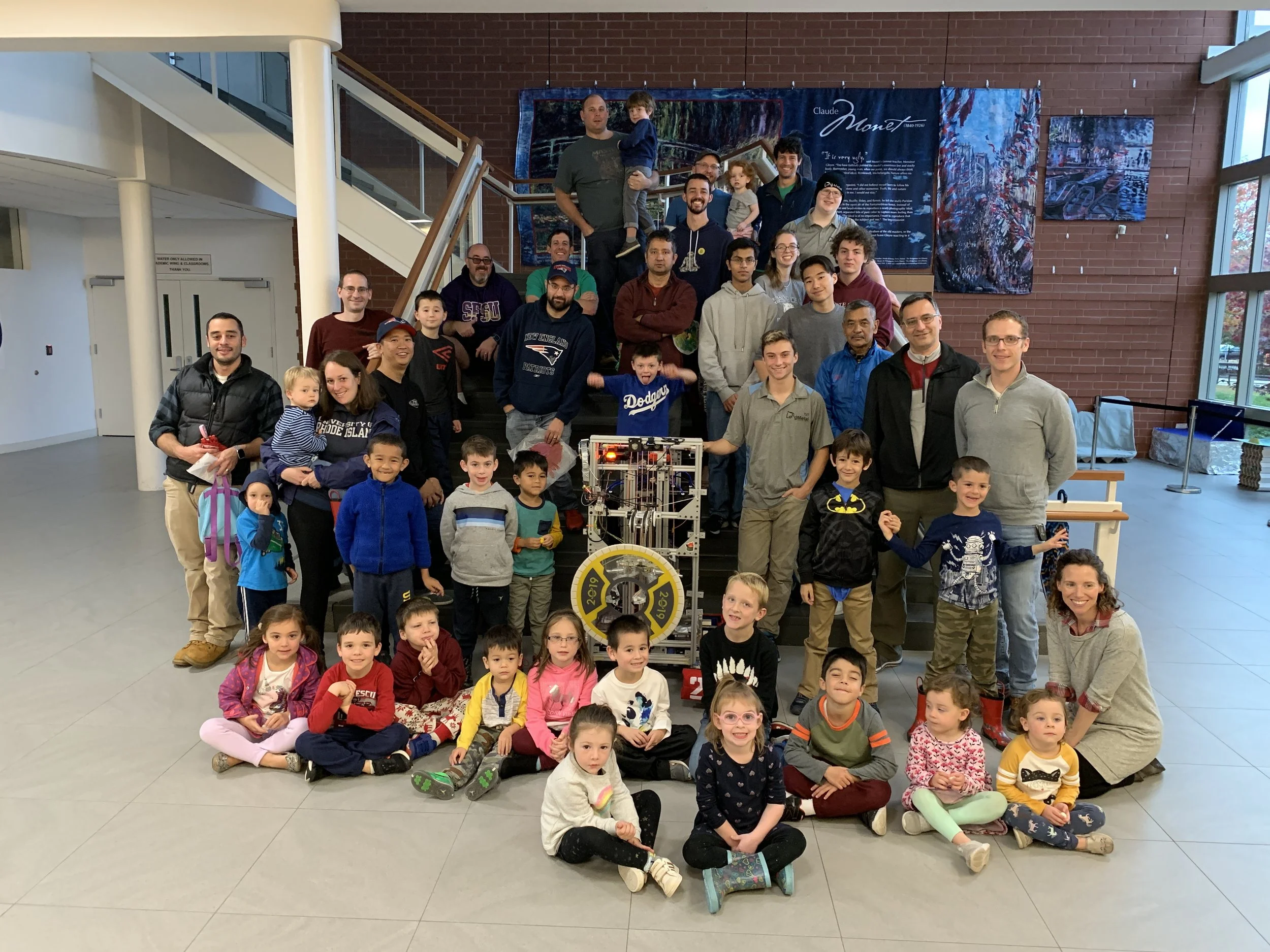 Group photo of children, adults, and a robot in a spacious indoor setting with a brick wall background and artwork.
