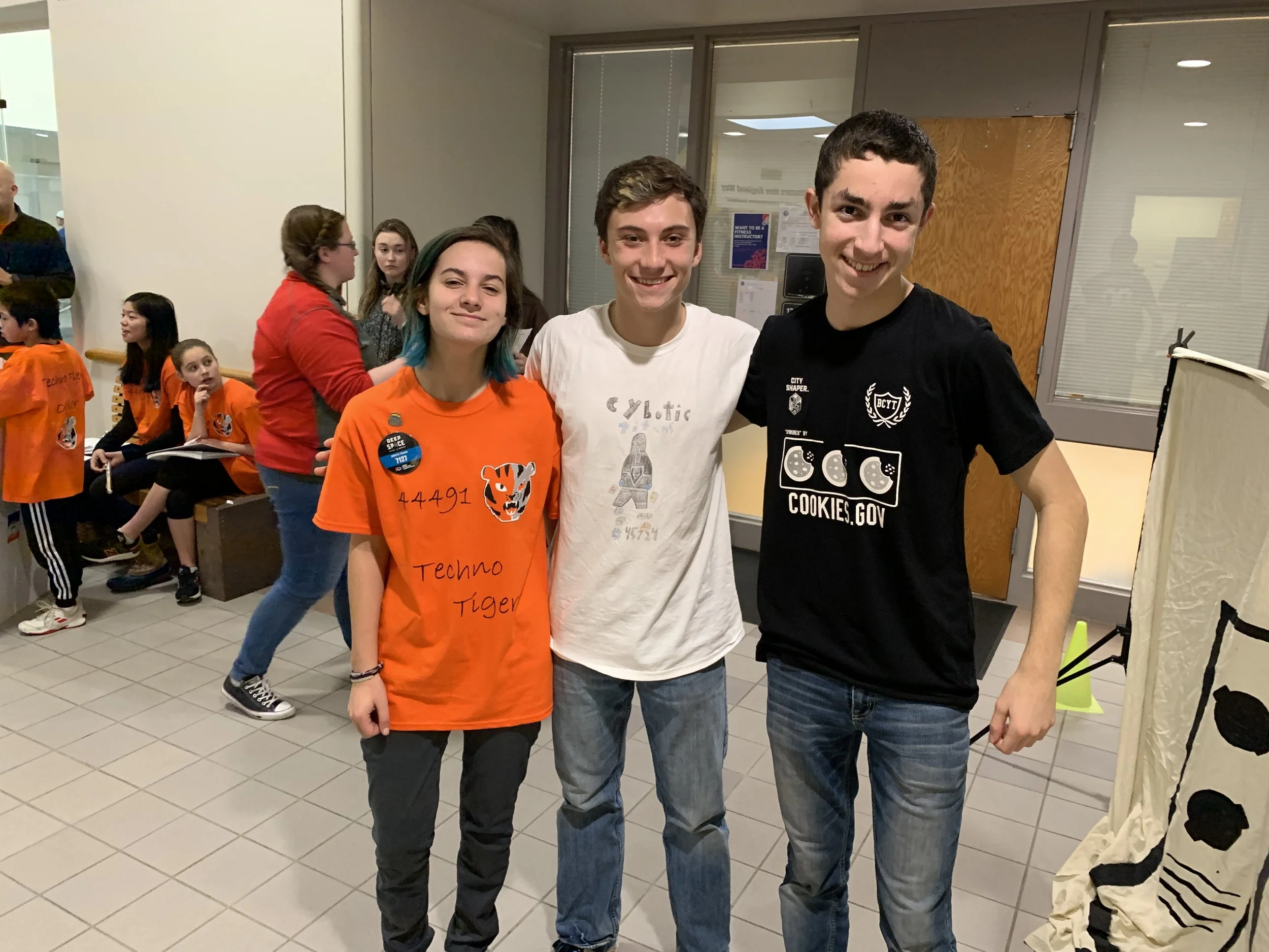 Three teenagers standing together indoors, smiling at the camera. The girl on the left is wearing an orange T-shirt with a tiger graphic and the words 'Techno Tiger,' the boy in the middle is wearing a white T-shirt with robot graphics, and the boy o
