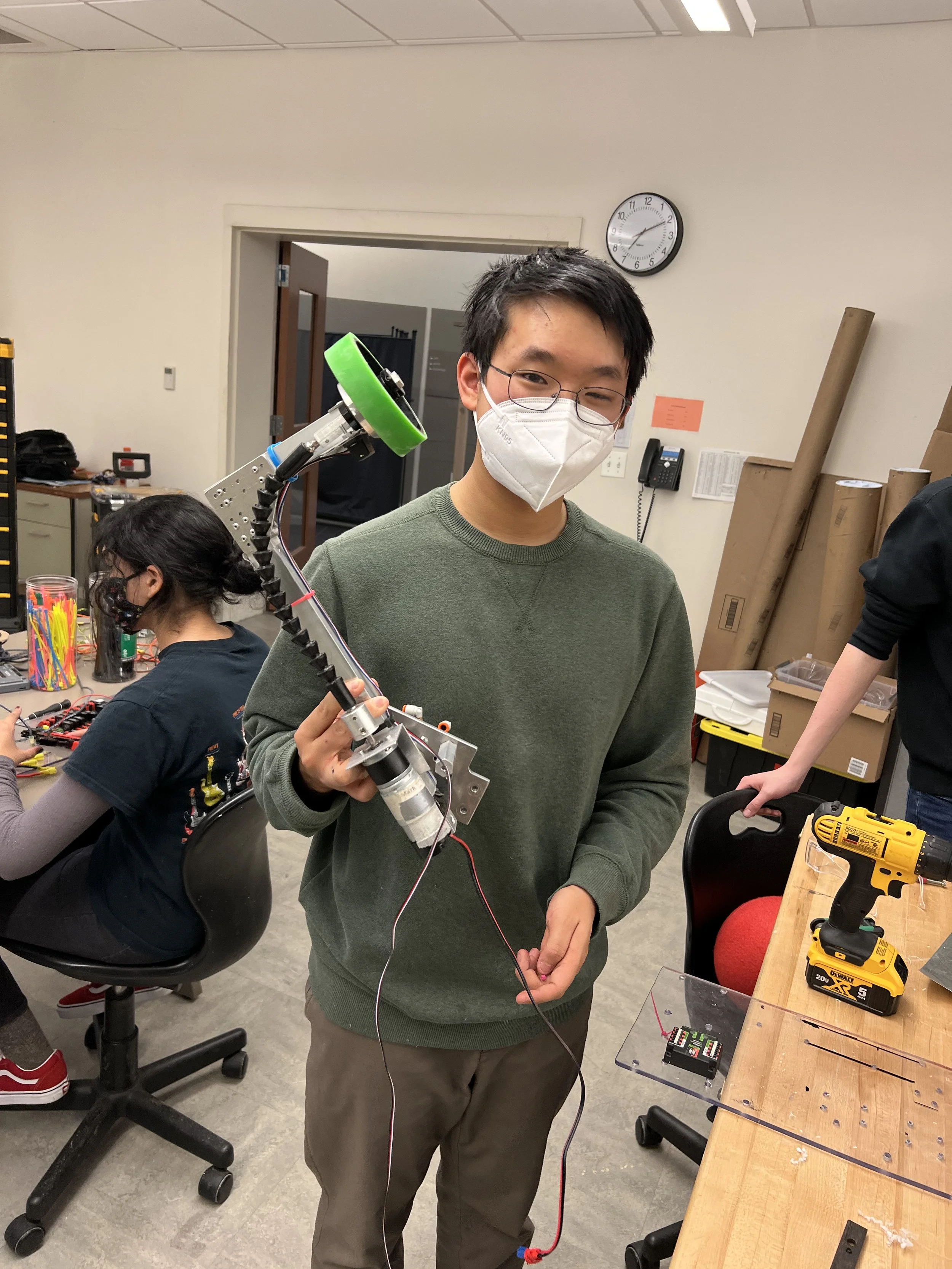 A young man wearing glasses and a face mask in a workshop, holding a robotic arm with a green wheel at its end. There are tools and other people working at tables around him.