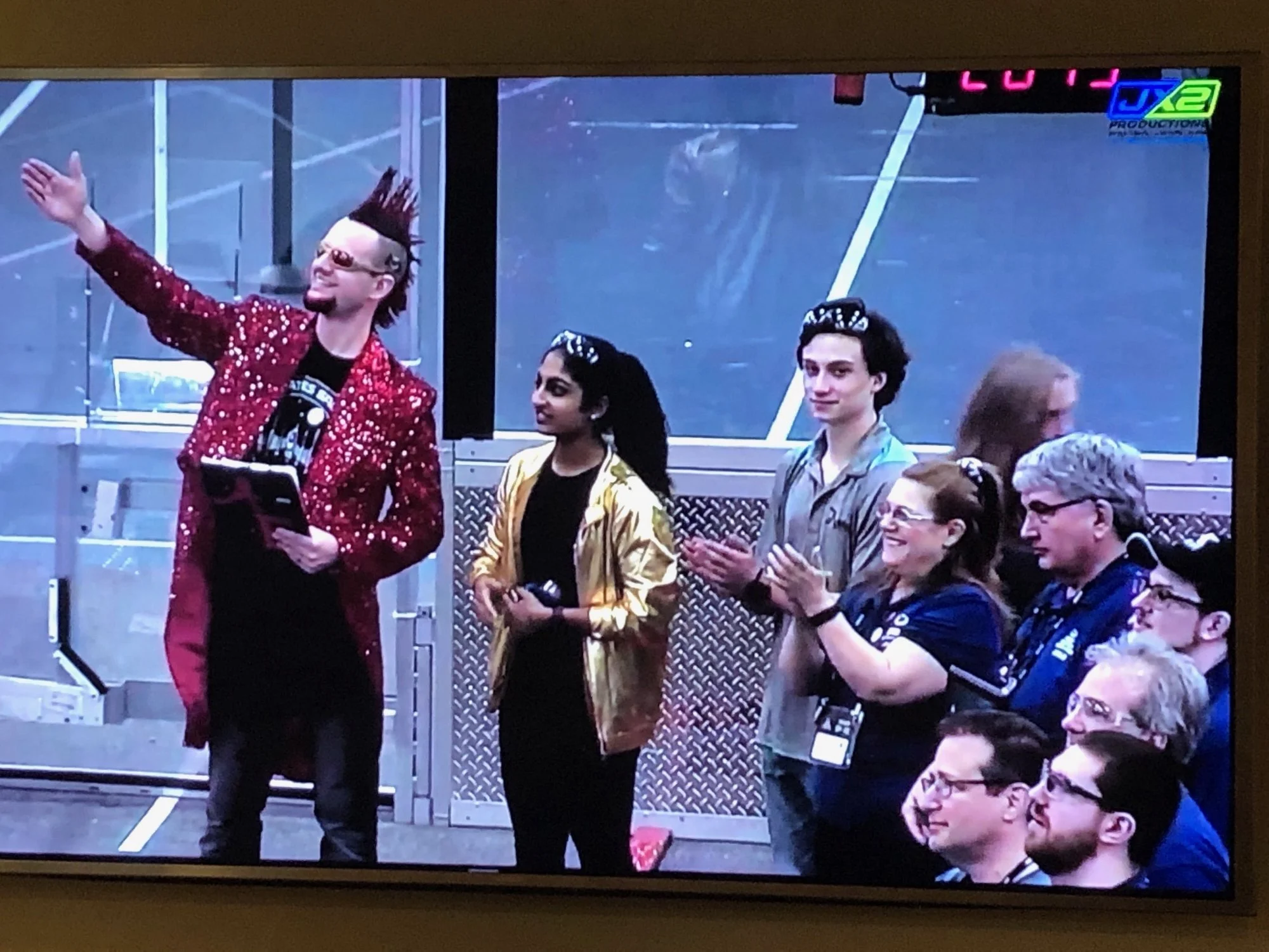 A group of people at a robotics competition, with one person in a red sequin blazer speaking into a microphone, while others watch and applaud.