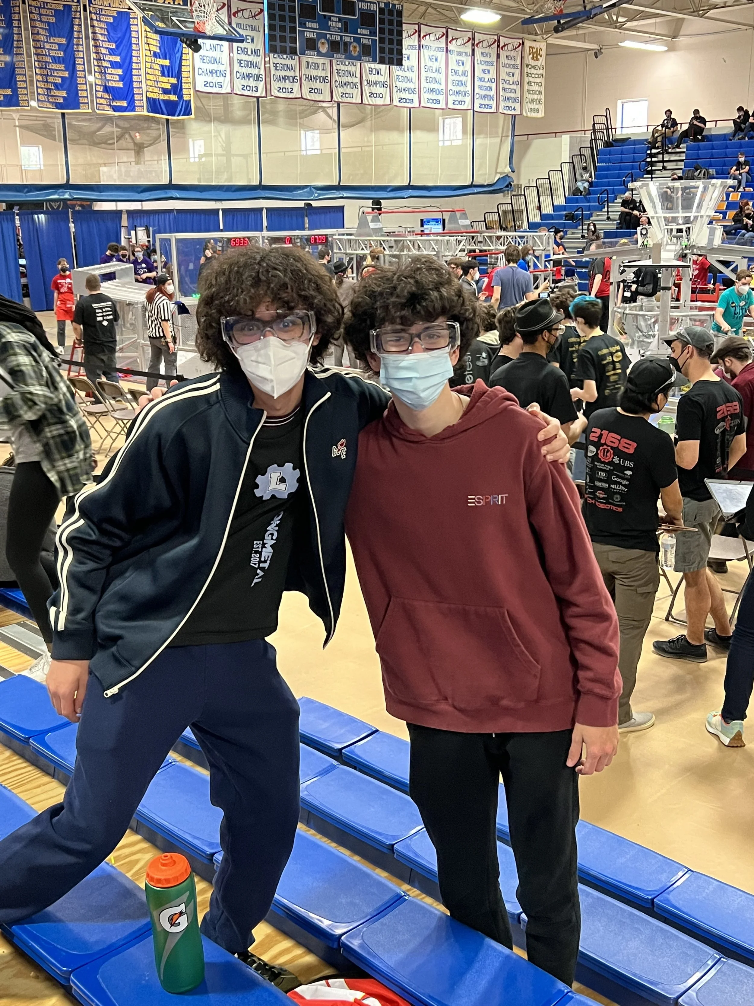 Two young men wearing face masks and glasses standing in a gymnasium during a robotics competition, with many people and robotics equipment in the background.