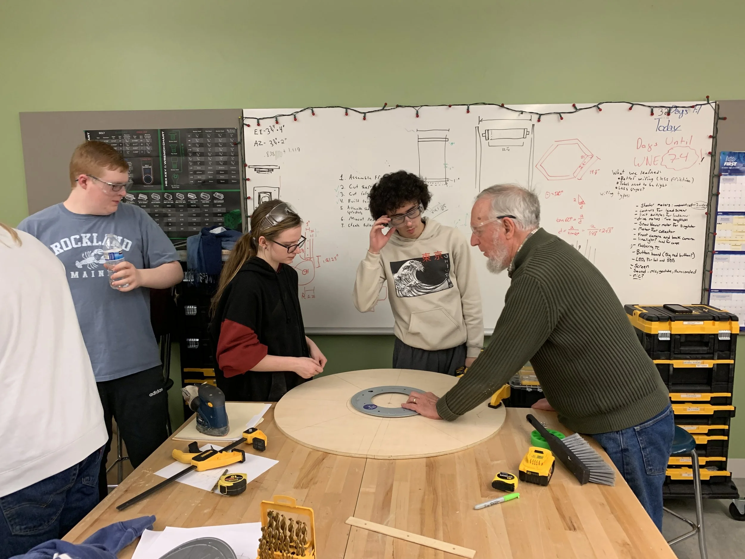 A group of students and an instructor working around a wooden table with tools, a protractor, and a circular piece, in a classroom with a whiteboard and tool storage in the background.