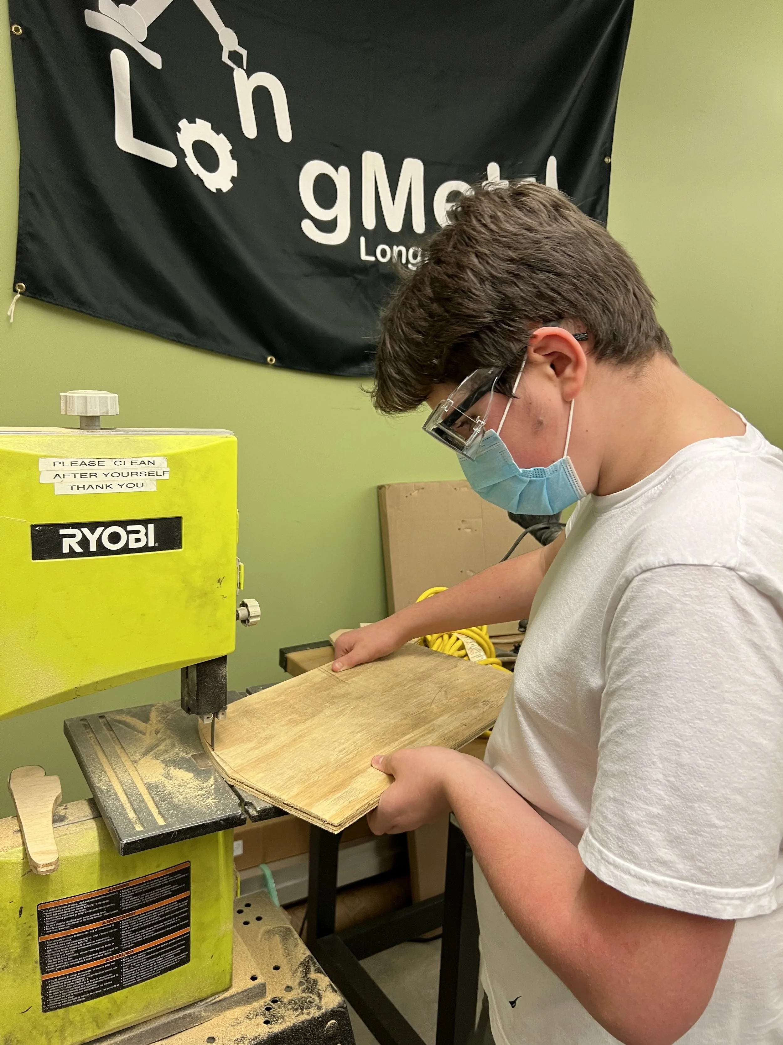 A young person wearing safety glasses and a face mask is operating a yellow Ryobi scroll saw, cutting a piece of wood in a woodworking workshop.