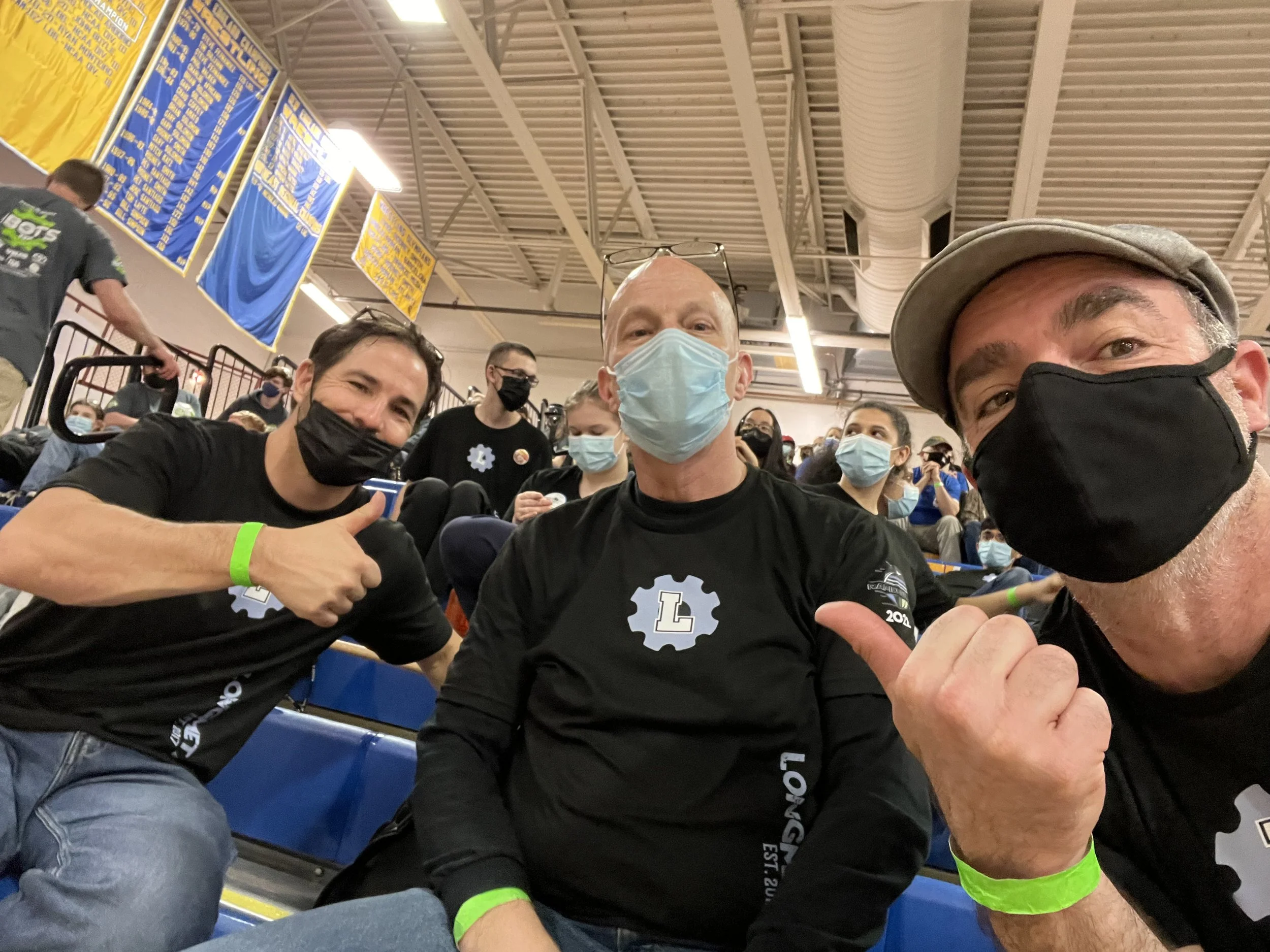 Three men wearing black masks and black long-sleeve shirts sitting in a gymnasium, with the man on the left giving a thumbs-up and the man on the right making a fist, surrounded by a crowd of masked people.