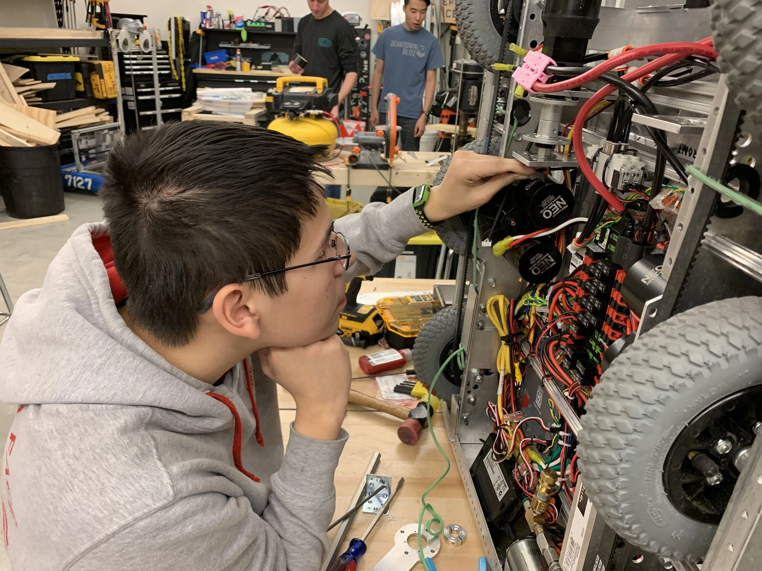 A young man working on a robotics project inside a workshop, with electronic components, wires, and tools around him, and two other people in the background working on similar projects.