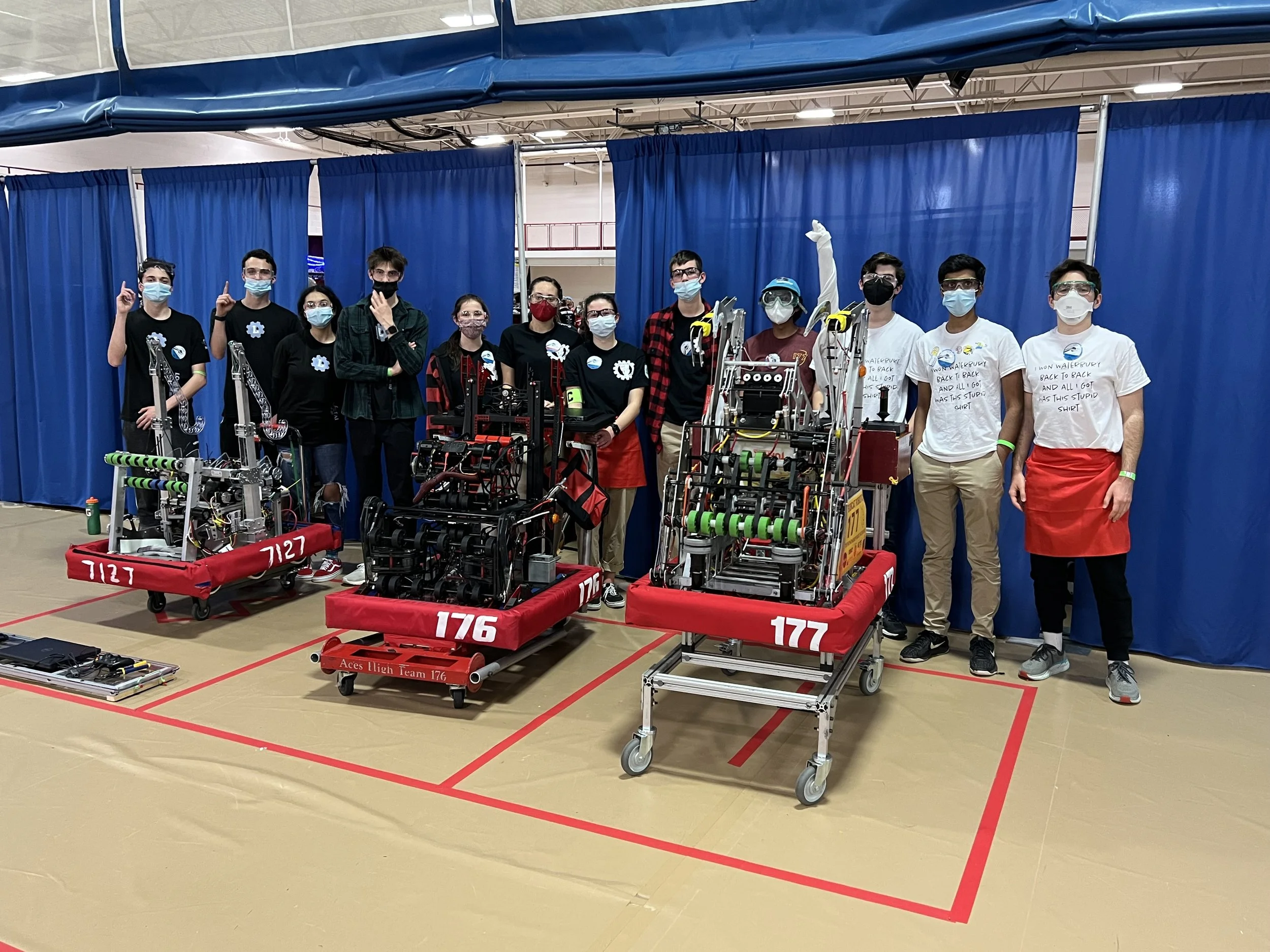 Team of students and robots at a robotics competition, standing behind their robots on a gymnasium floor with blue curtains.