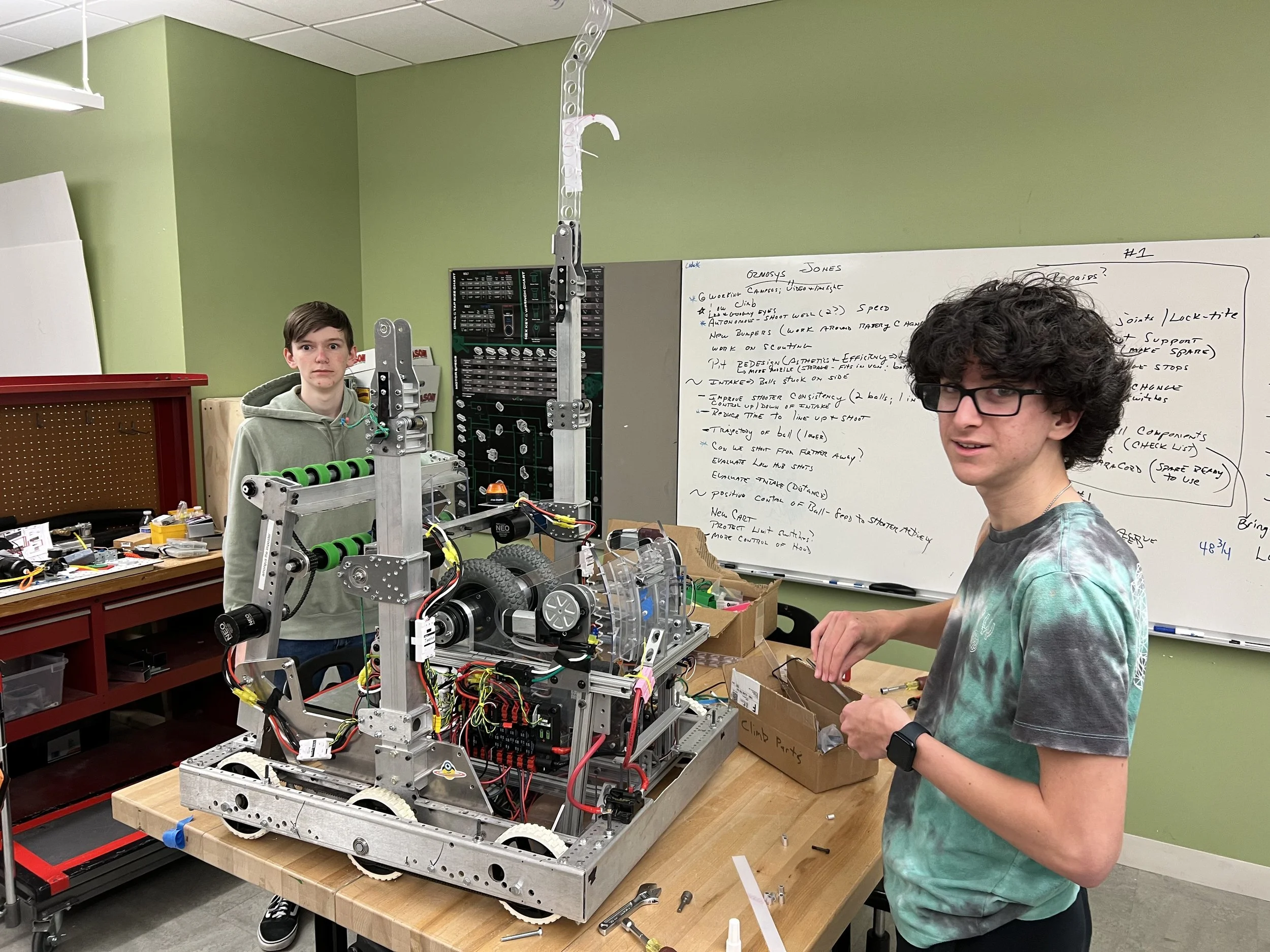 Two young men working on a robot in a workshop, with a whiteboard filled with notes behind them.