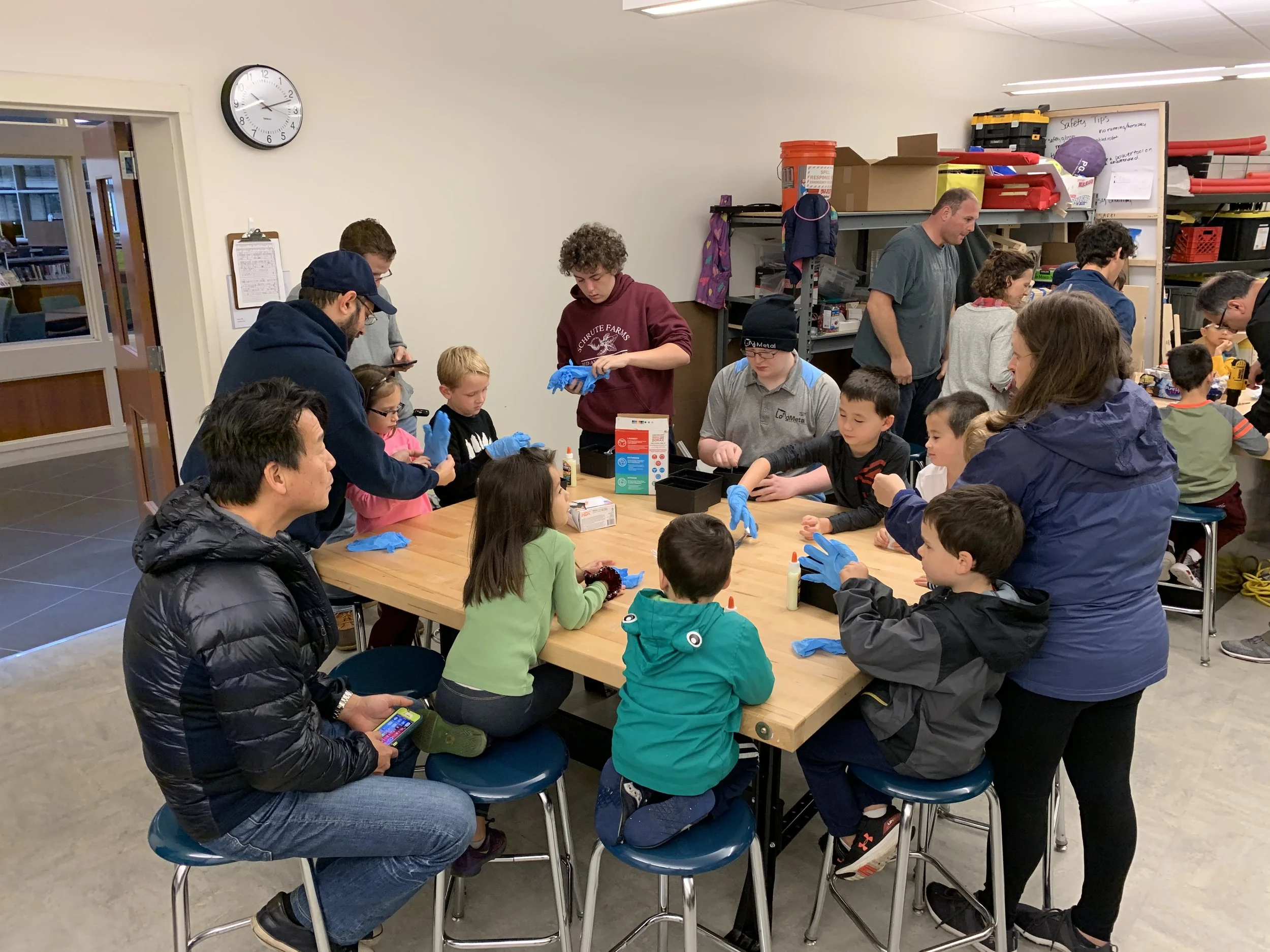 A group of children and adults gathered around a table in a workshop, making arts and crafts projects with glue and supplies, with shelving and a whiteboard in the background.