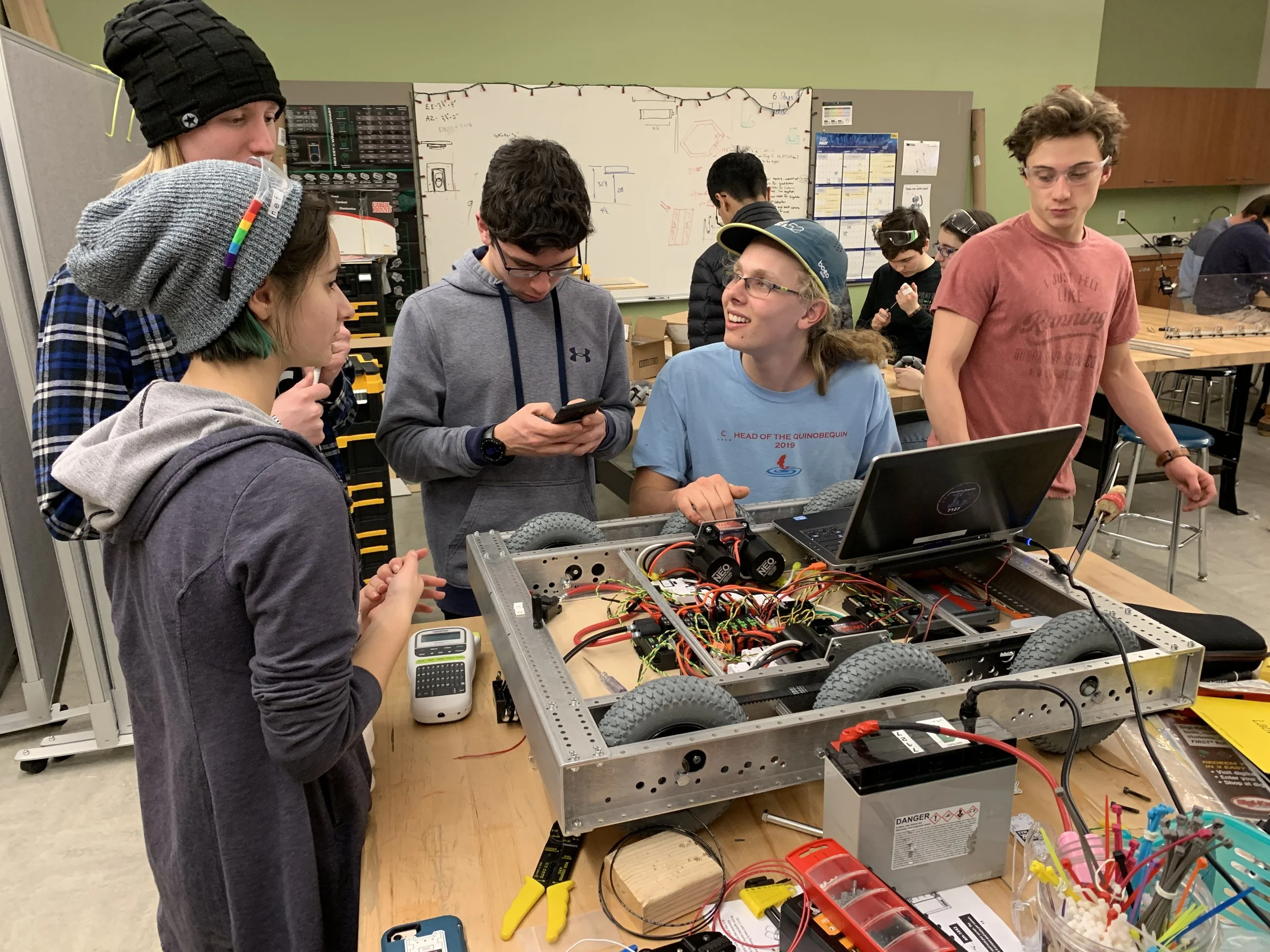 Group of students working on a robotics project in a classroom workshop, with some students wearing protective gear and using tools like a calculator, smartphone, and laptop.