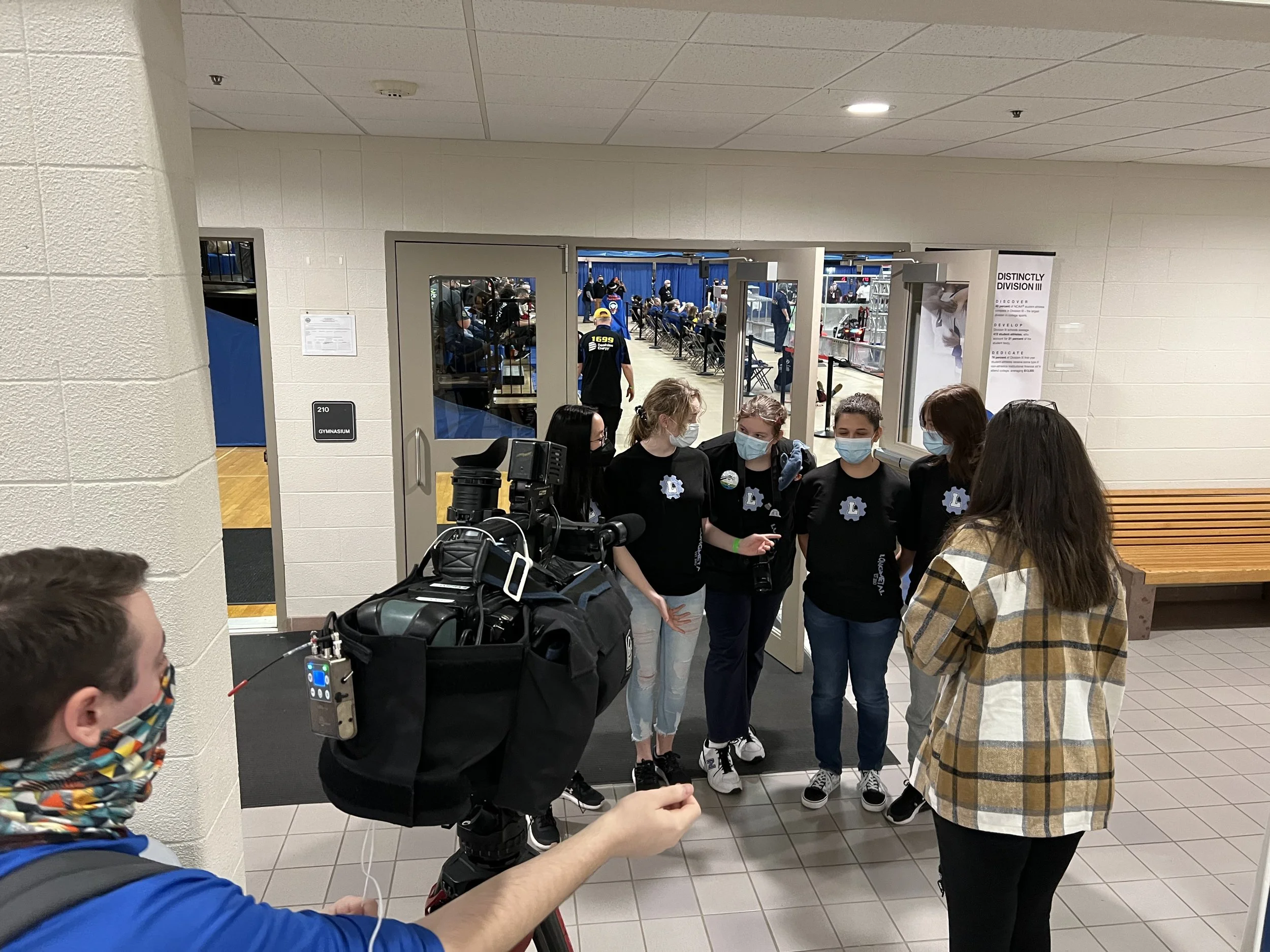 A group of young women, wearing masks and black t-shirts, being interviewed by a person with long hair wearing a plaid jacket in a school hallway near door 210 and a gymnasium.