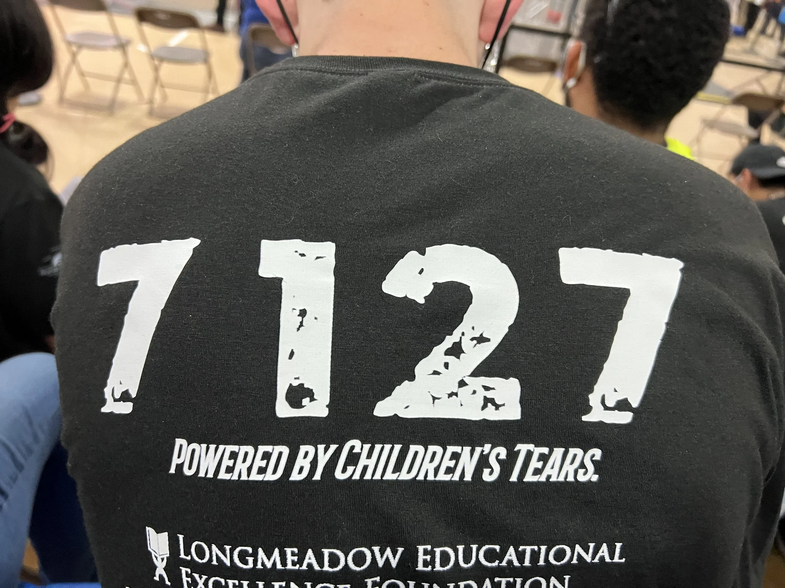 Back of a person wearing a black T-shirt with the words '7.1.21 Powered by Children's Tears' and 'Longmeadow Educational Foundation' printed on it, sitting at an indoor event with chairs and other people visible in the background.