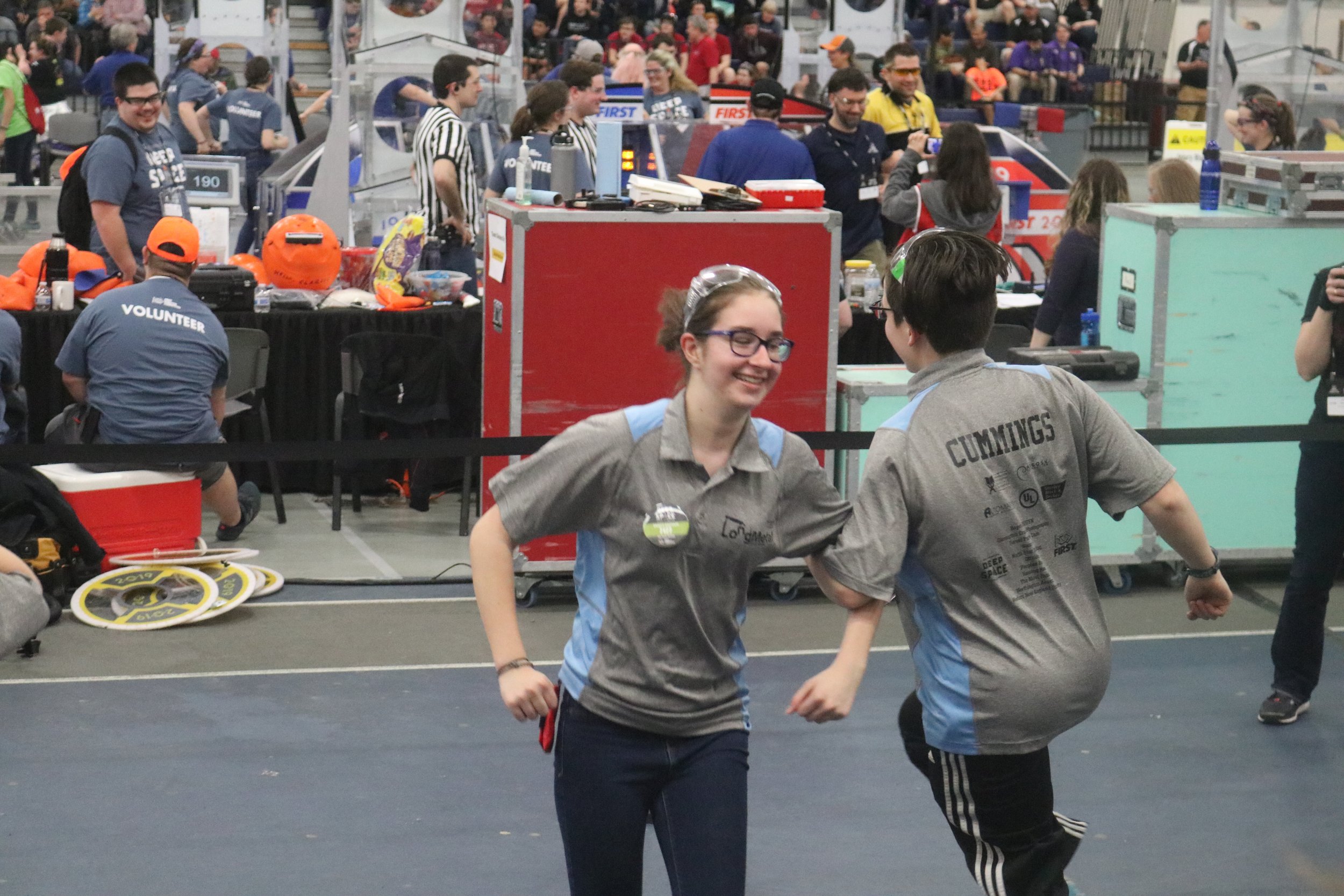 Two young women at a robotics competition, smiling and holding hands, with other participants and team tables in the background.