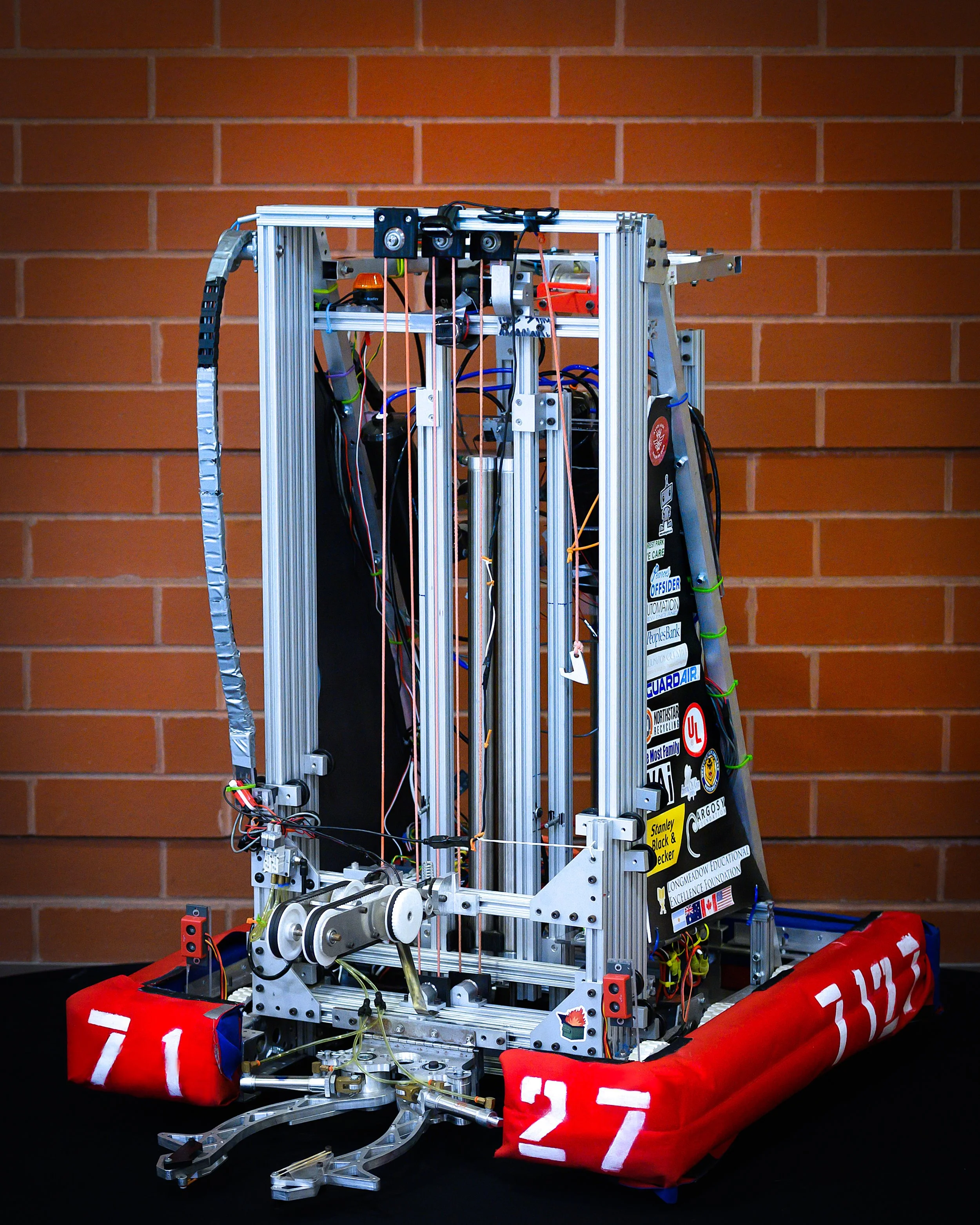 A robot made of metal and wires with racing numbers 71 and 27 on red protective pads, positioned against a brick wall.